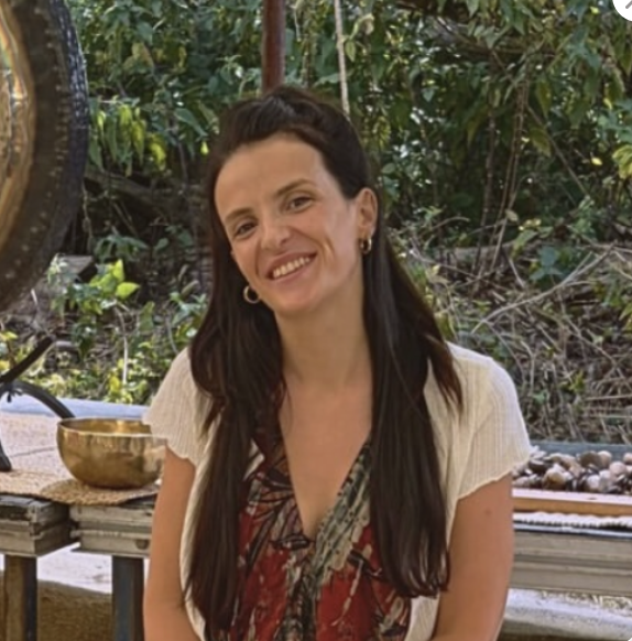 A woman with long dark hair and earrings smiling outdoors surrounded by greenery, with a musical instrument and bowl in the background.