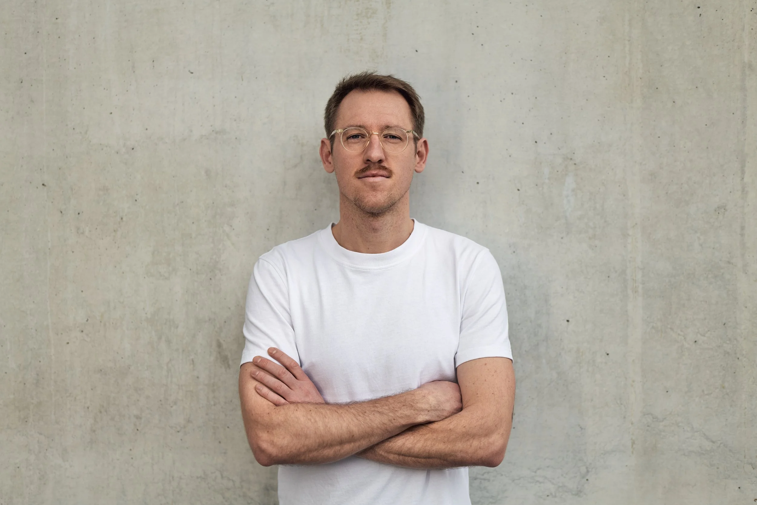 Junger Mann trägt weiße T-Shirt, steht vor einer grauen Betonwand, Arme verschränkt, schaut ernst in die Kamera, trägt Brille.