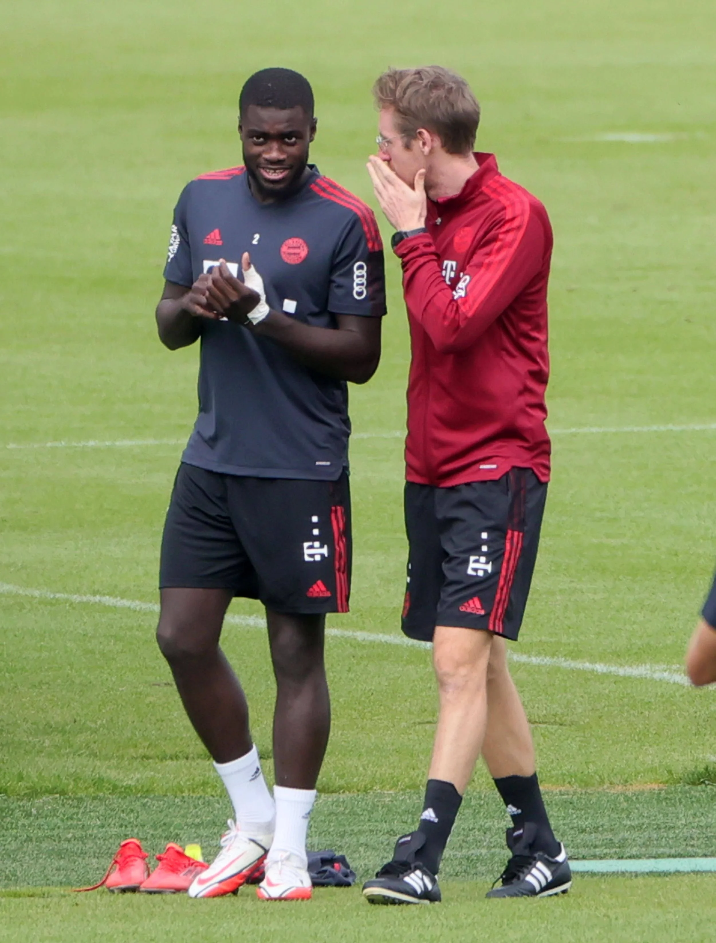 Zwei Fußballspieler im Gespräch auf dem Spielfeld, einer trägt schwarzen Trainingsanzug und der andere rote, beide mit Adidas-Logo, im Hintergrund grüne Spielfläche.