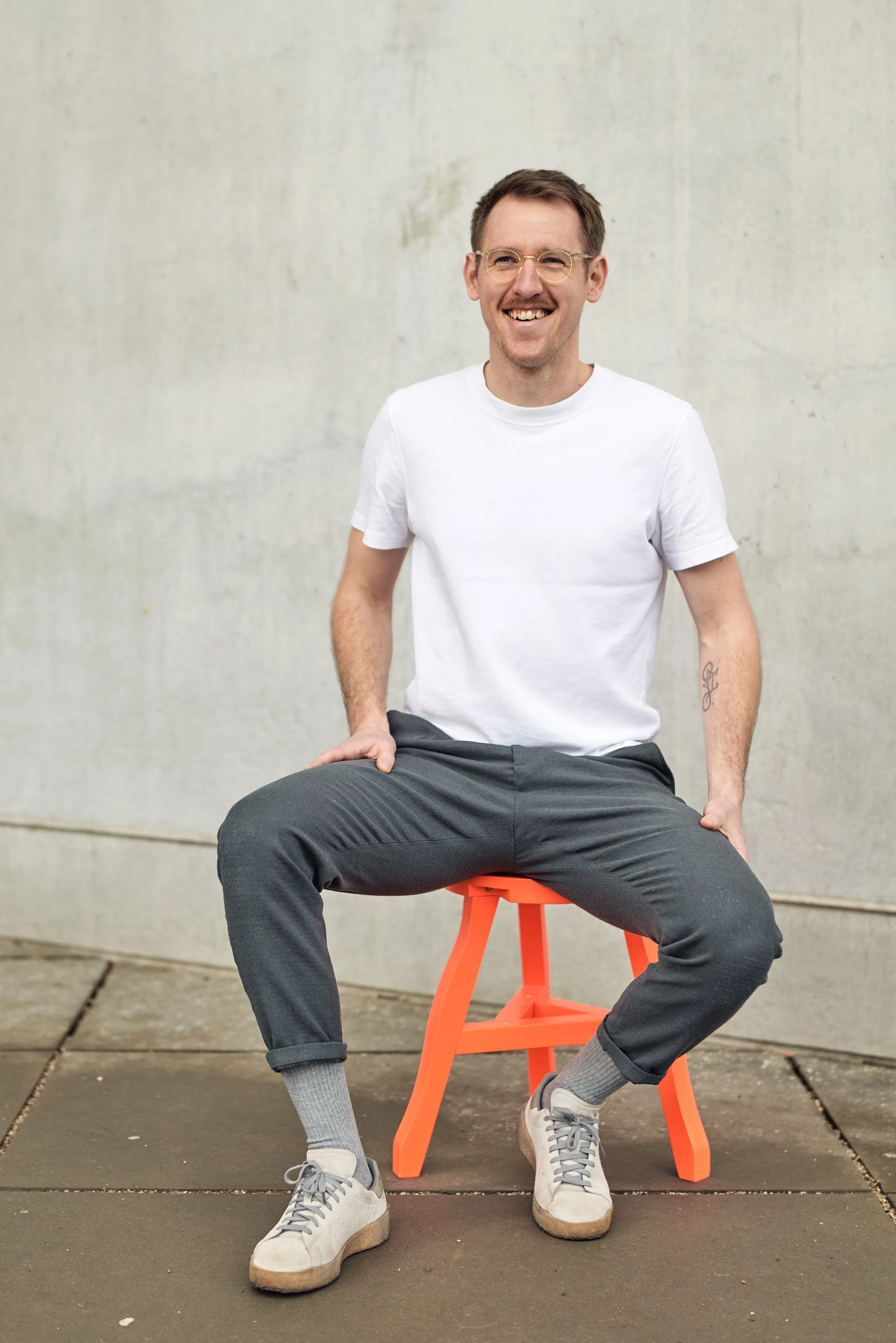 Ein lachender Mann in weißen T-Shirt und grauen Hosen sitzt auf einem orangefarbenen Hocker vor einer hellen Wand.