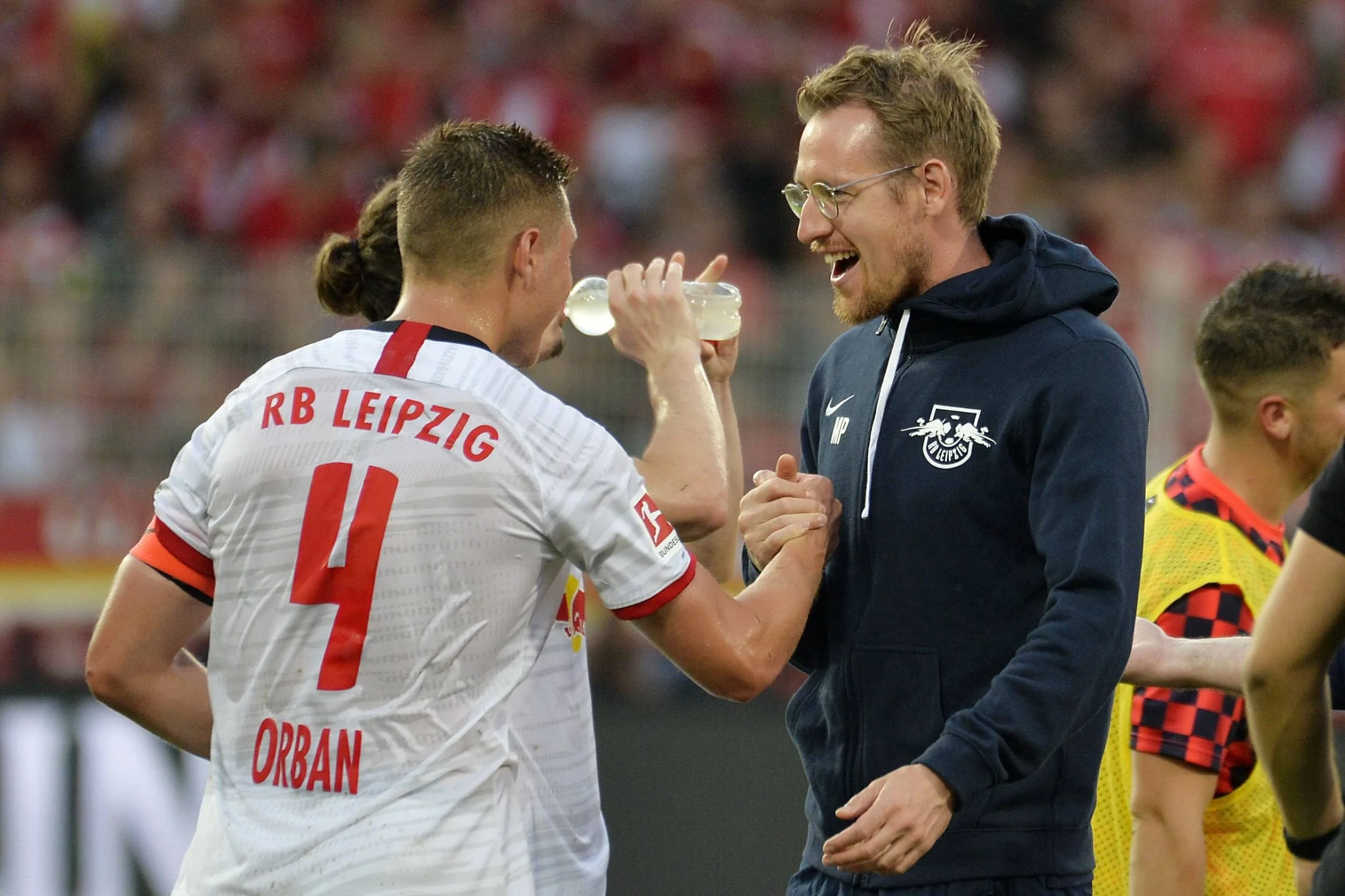 Fußballspieler in einem weißen Trikot mit Nummer 4 und Namen ORBAN, sowie einem Trainer im blauen Hoodie, die sich fröhlich die Hand geben auf dem Spielfeld bei einem Fußballspiel.