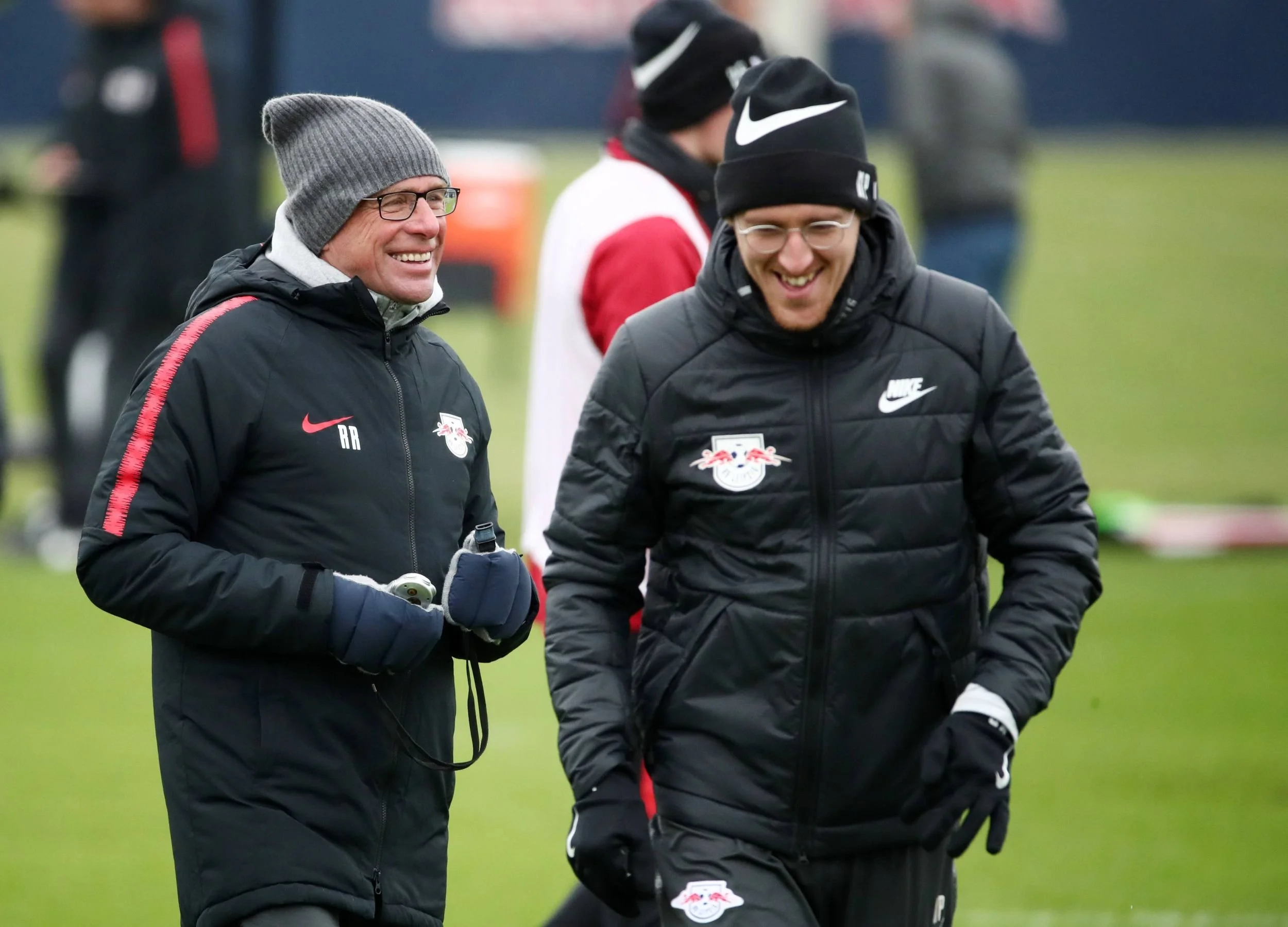 Zwei Männer in Winterkleidung und Brillen, die lachend auf einem Fußballplatz laufen, mit dem RB Leipzig Logo auf ihren Jacken.