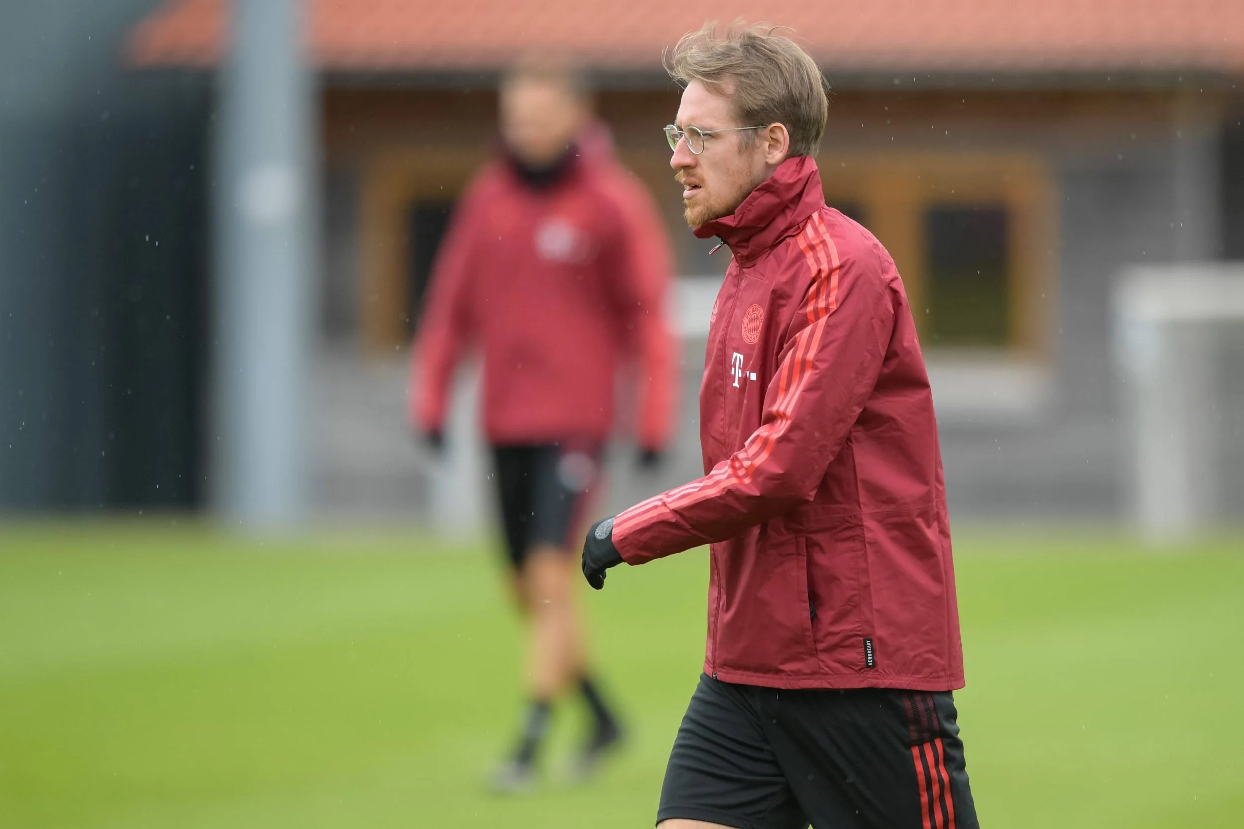 Ein Mann in roten Sportjacken und schwarzen Hosen, möglicherweise ein Trainer, steht auf einem Sportplatz bei einem Fußballtraining.