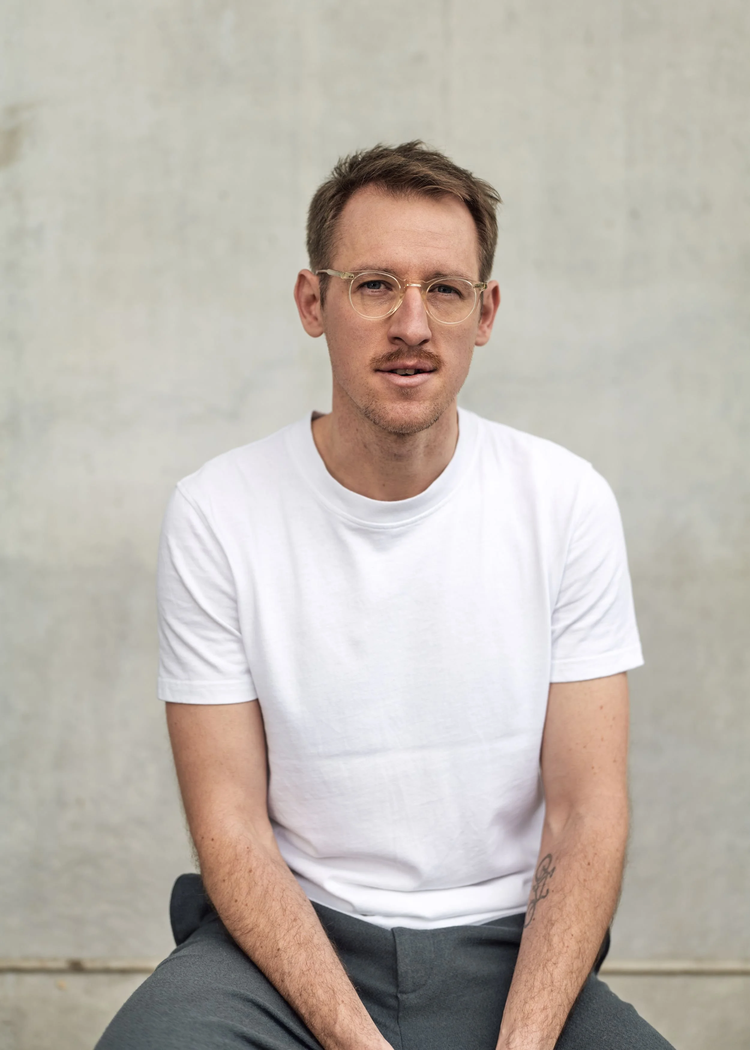 Ein Mann mit Brille, kurzen braunen Haaren, trägt ein weißes T-Shirt und graue Hosen, sitzt vor einer hellen Wand.