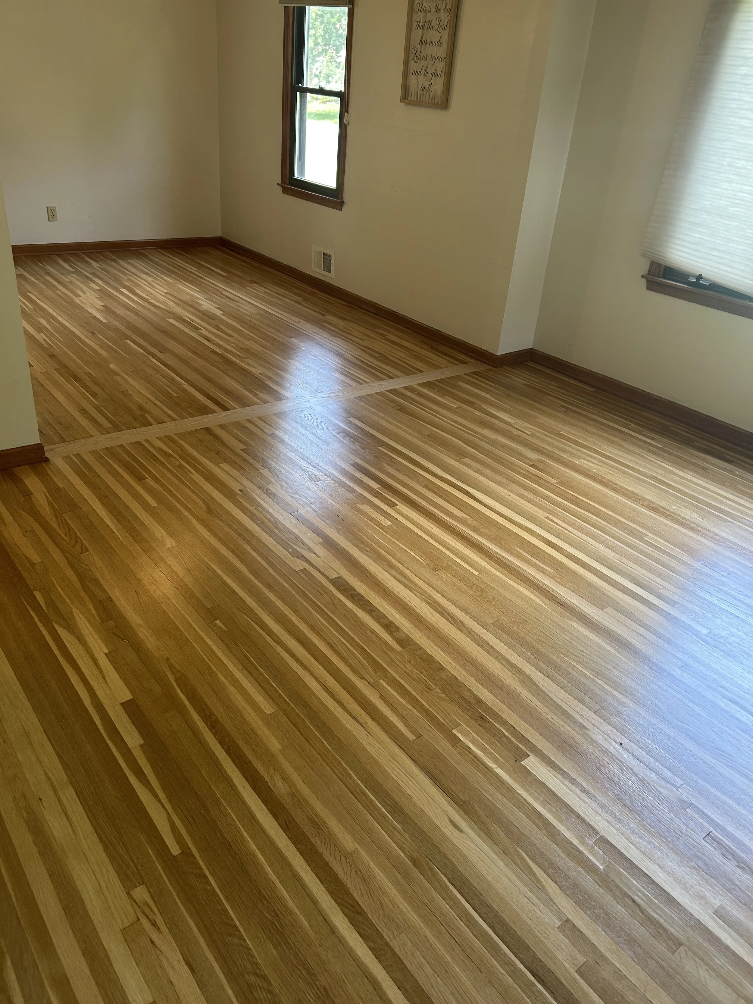 Empty room with hardwood floors, white walls, two windows with blinds, and a framed quote on the wall.