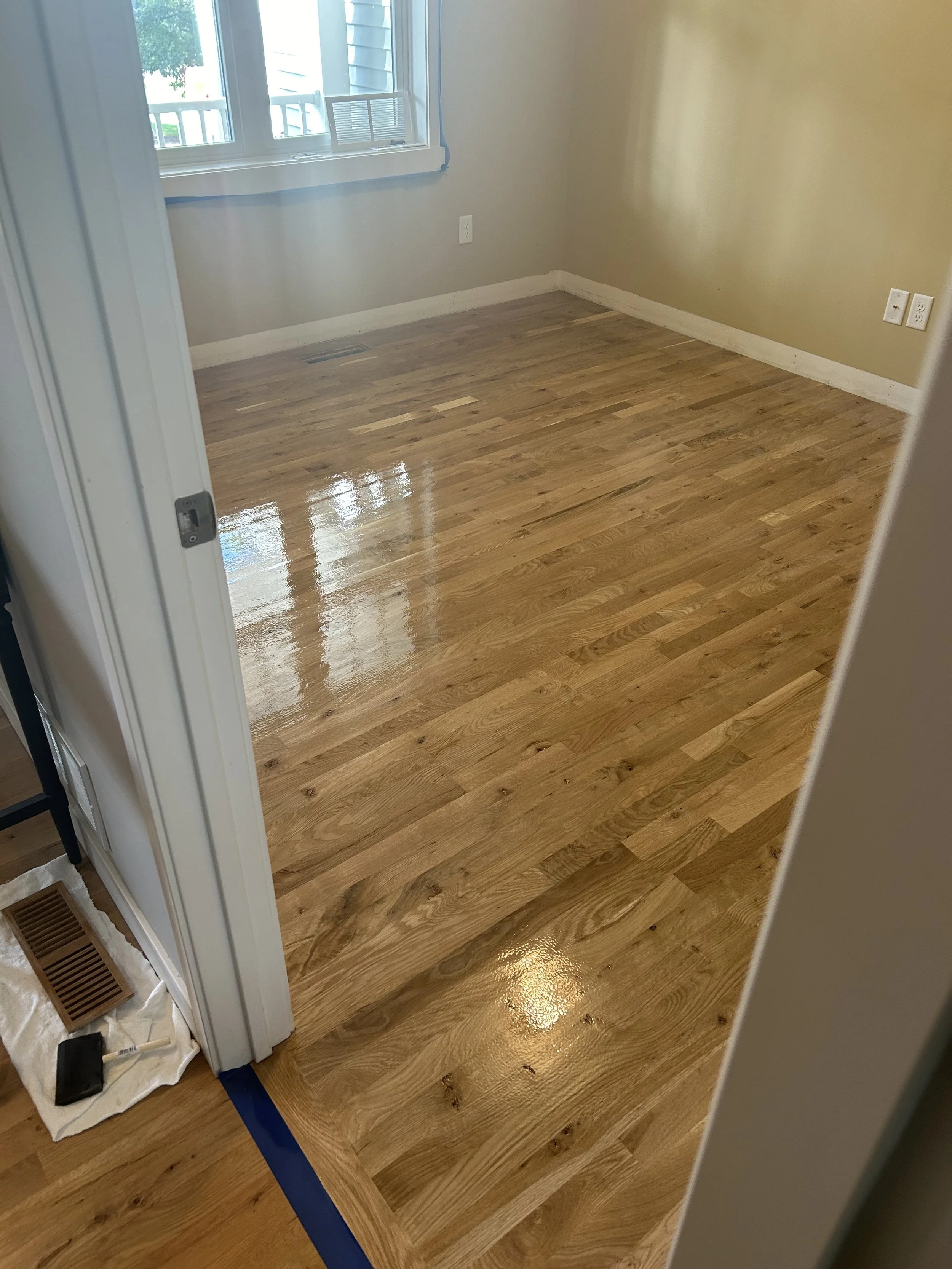 A room with freshly polished hardwood floors, a window with sunlight coming through, beige walls, and electrical outlets on the wall.