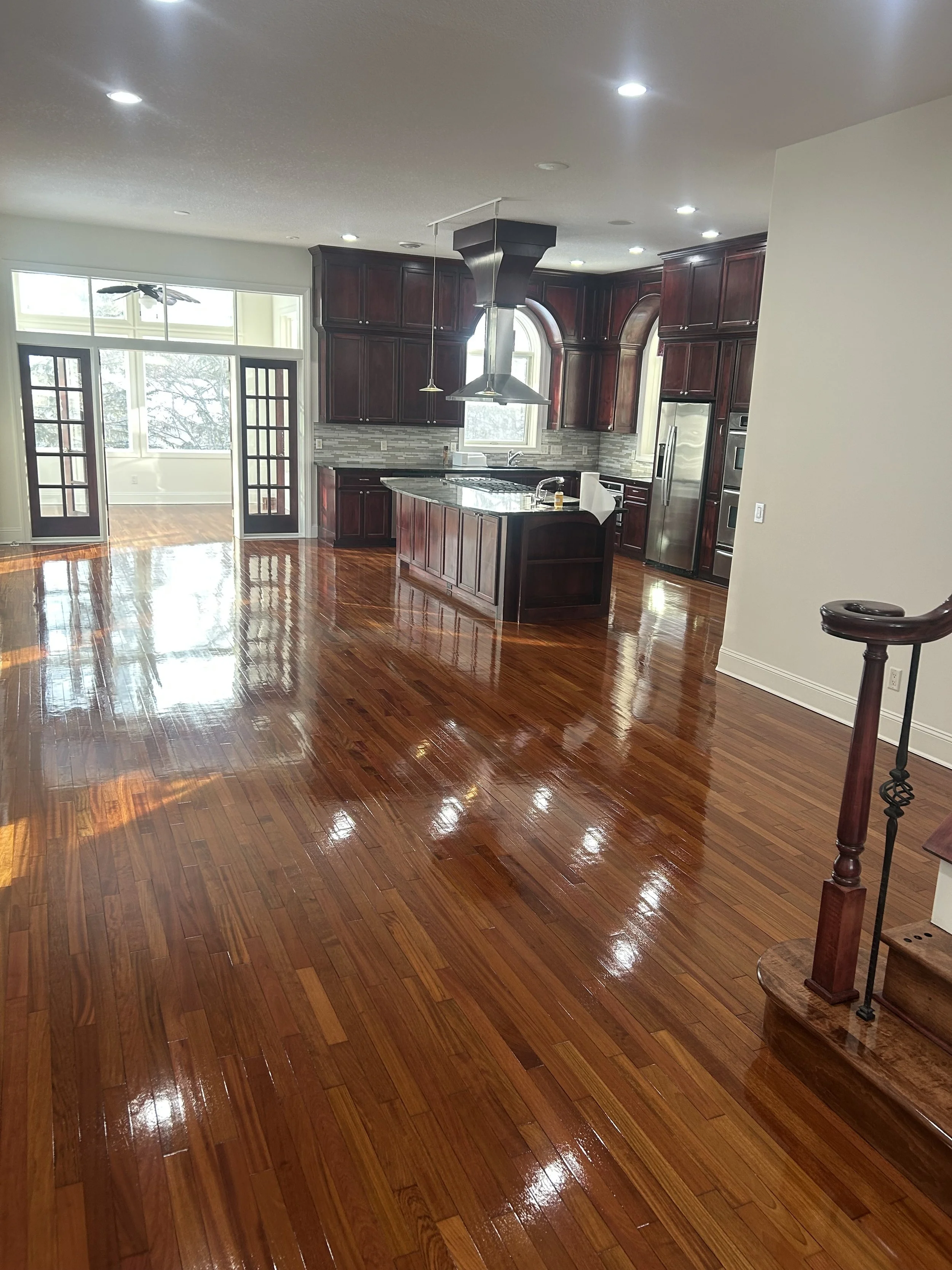 Bright open kitchen with dark wood cabinets, granite island, stainless steel appliances, large windows, and high-gloss hardwood floors.