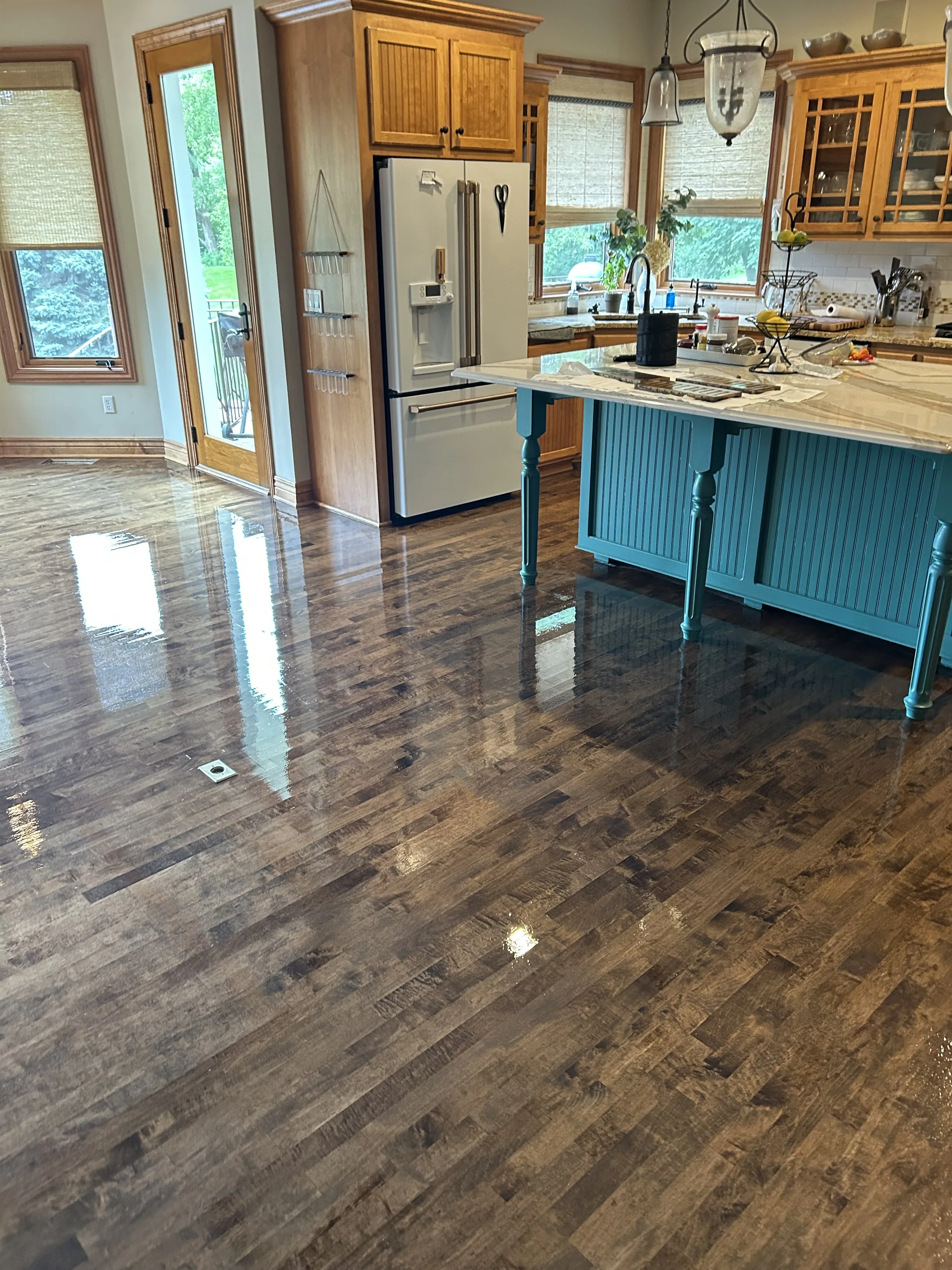 Recently cleaned and polished hardwood kitchen floor with shiny, reflective surface, showing part of a kitchen with wooden cabinets, a teal island, and a refrigerator.
