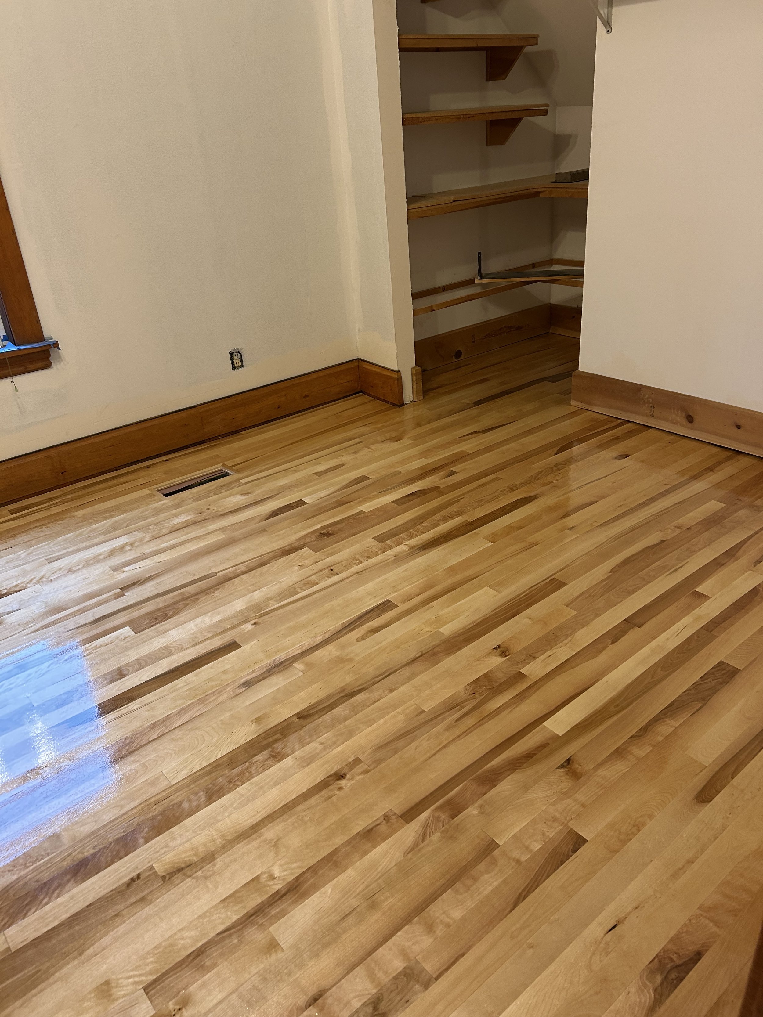 Clean, polished hardwood floor in a room with beige walls, a small window, and an open closet with shelves.