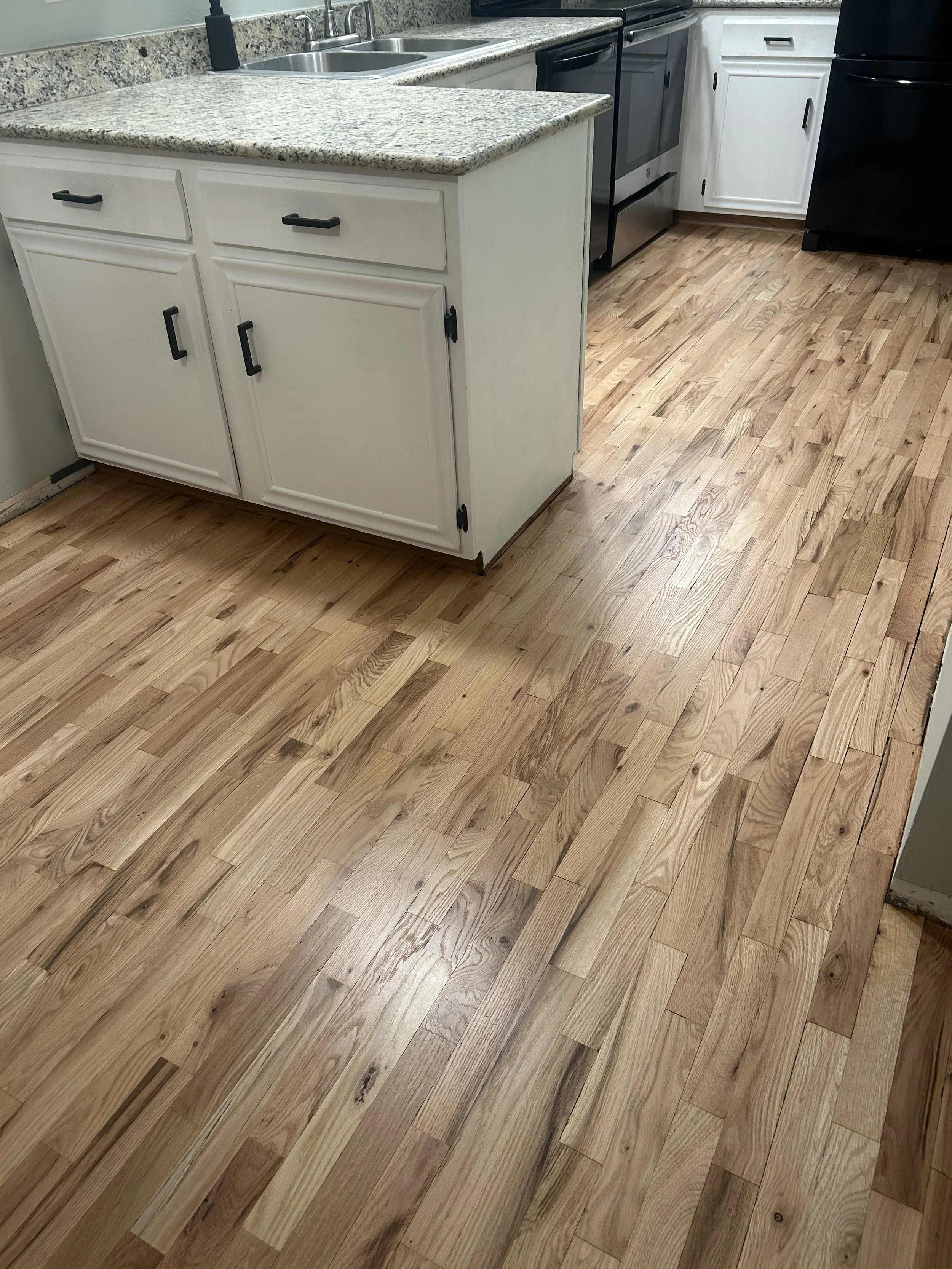 Wooden kitchen floor with light and dark wood planks, white cabinet with black handle, granite countertop, and stainless steel sink. Black appliances are visible in the background.