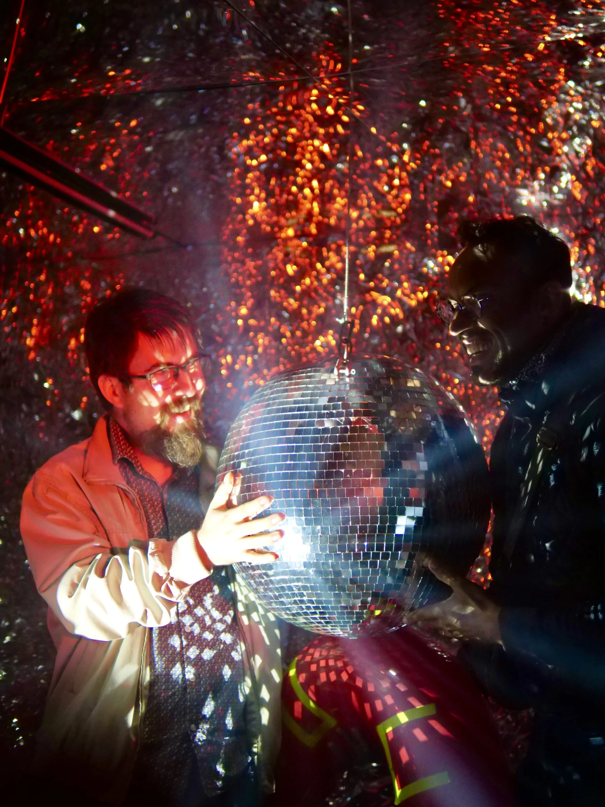 Deux hommes souriants touchent un grand ballon de discobole dans un décor lumineux avec des reflections et des lumières rouges et blanches, créant un effet de discothèque.