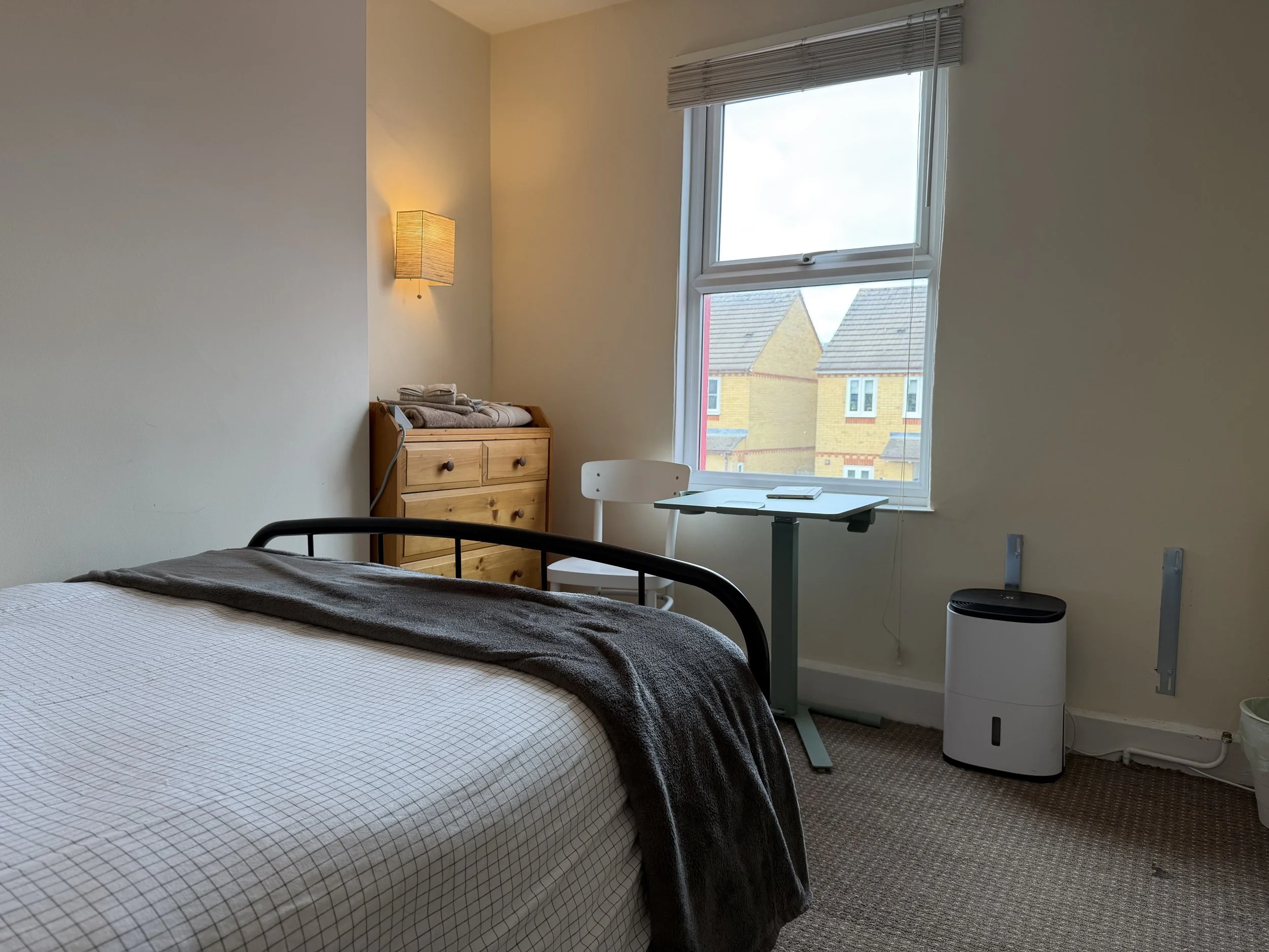 A bedroom with a bed, wooden dresser, small desk with a chair, window with a view of neighboring houses, wall-mounted light, and a humidifier.