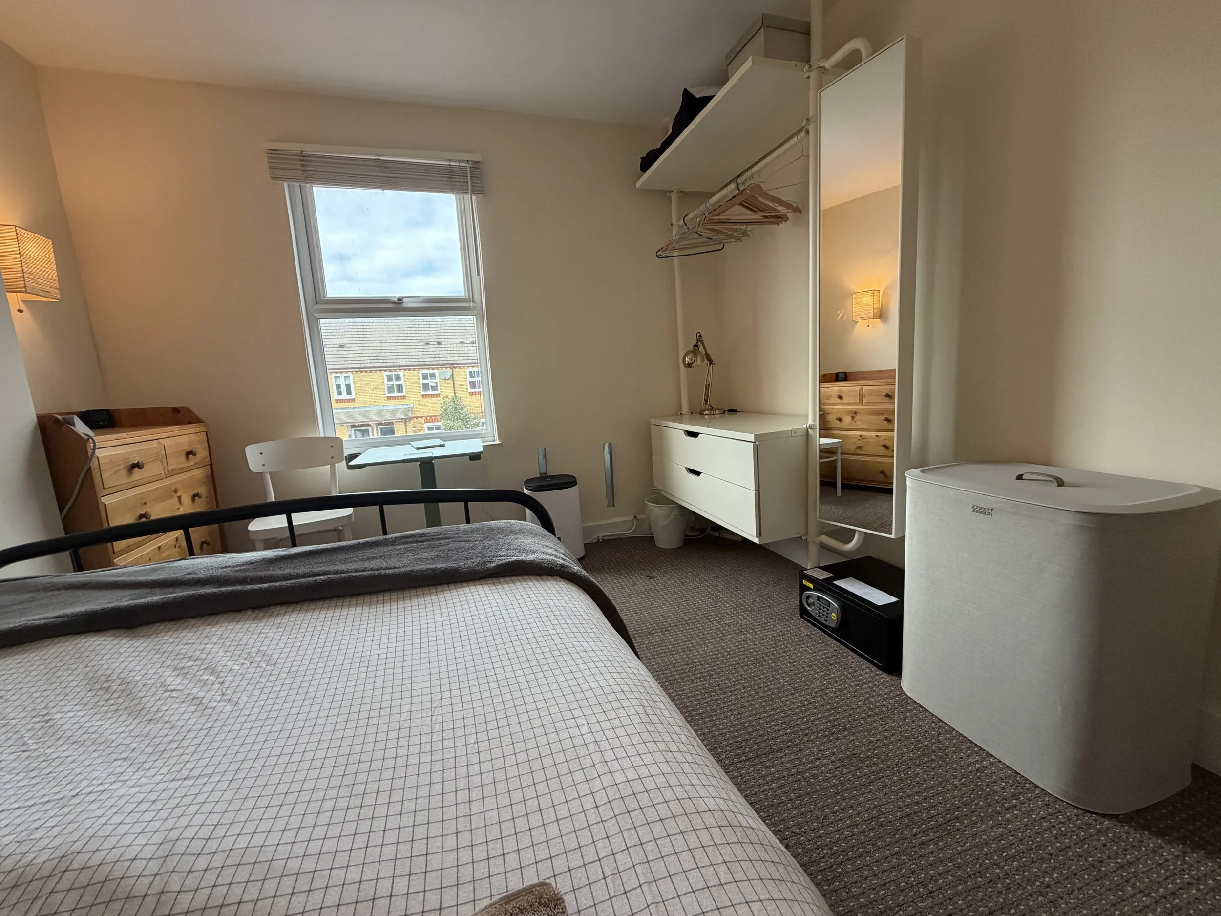 A bedroom with a bed, a window with view of houses outside, a white chair, a dresser, a mirror, and a mini safe.