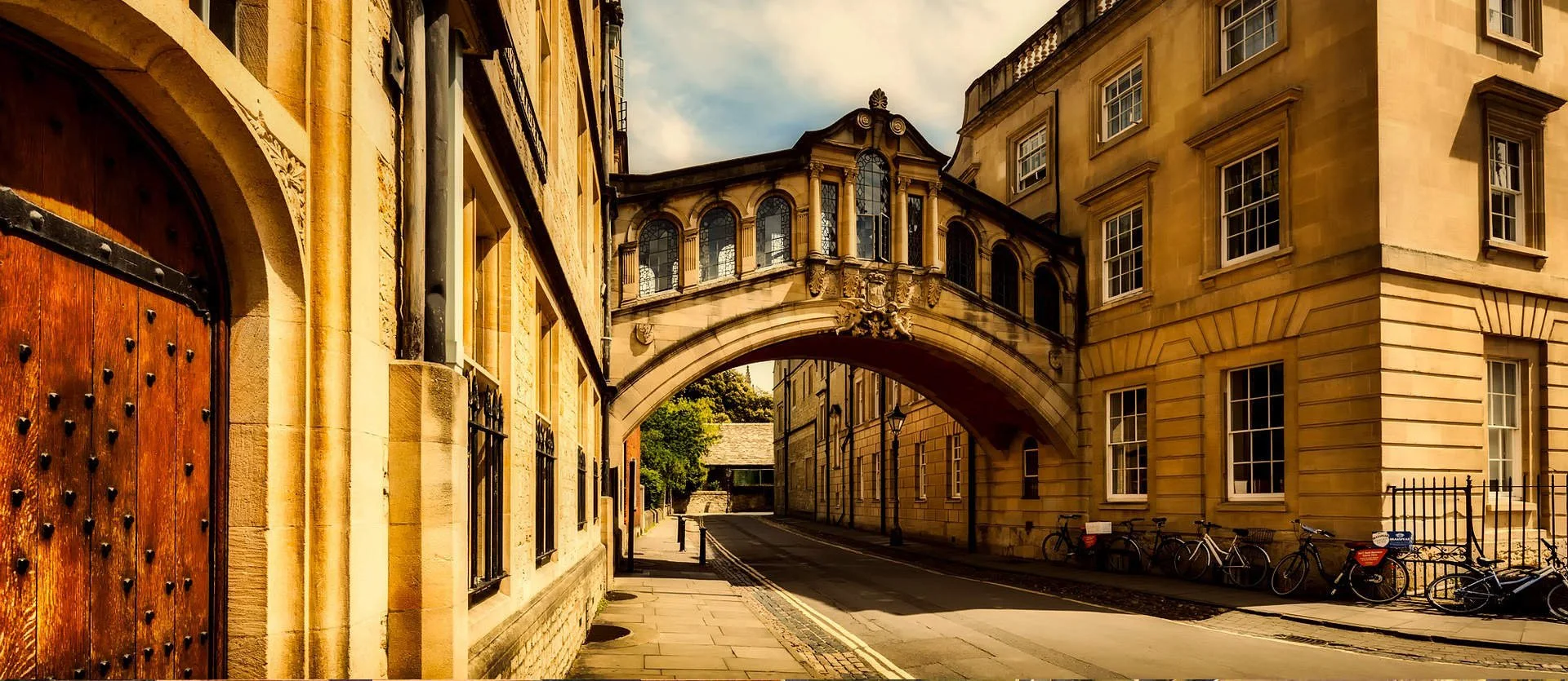 Historical street view with stone buildings, decorative bridge connecting buildings, bicycles parked along sidewalk, and cobblestone street.