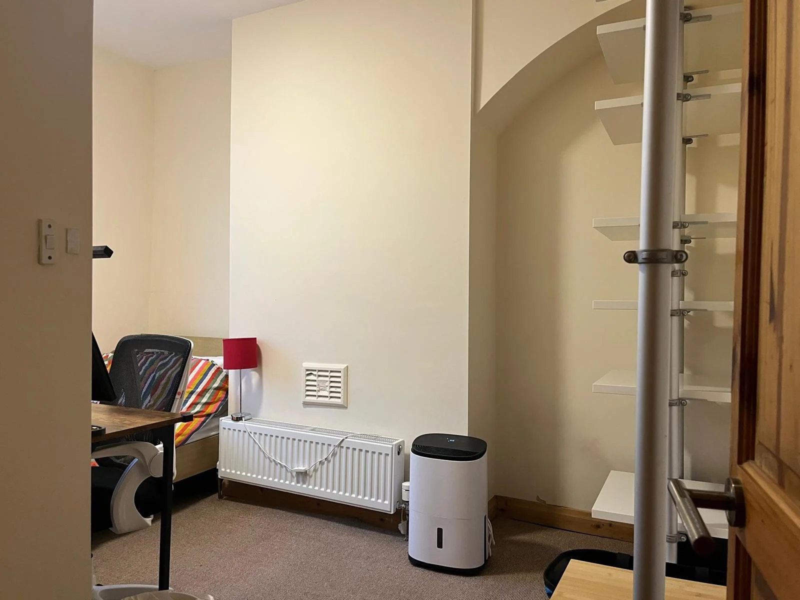 A room with beige walls, a bed with striped bedding, a black office chair, a small red lamp, a white radiator, a white air purifier, and a white shelving unit with metal supports.