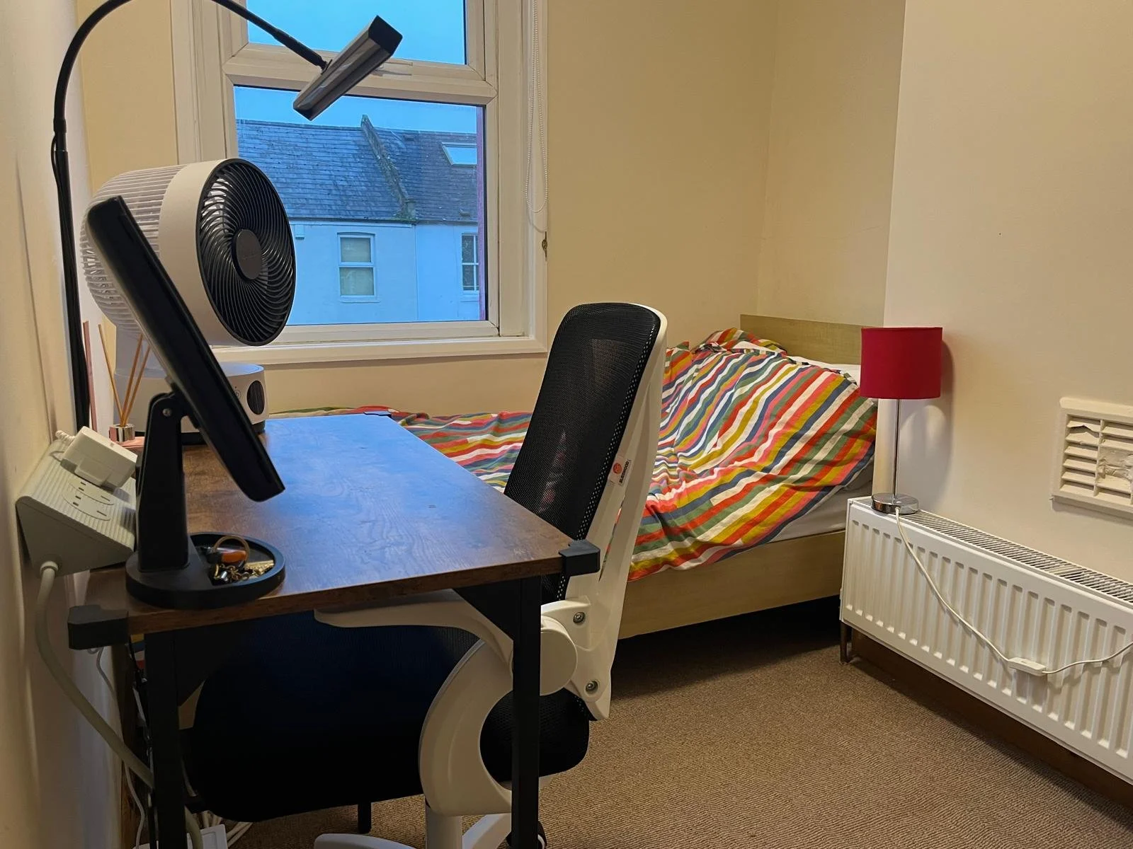 A bedroom with a wooden bed covered in a colorful striped duvet, a red bedside lamp, a black desk chair, a wooden desk with a black fan, a computer monitor, a white newsletter, a window showing neighboring houses, and a beige carpet.