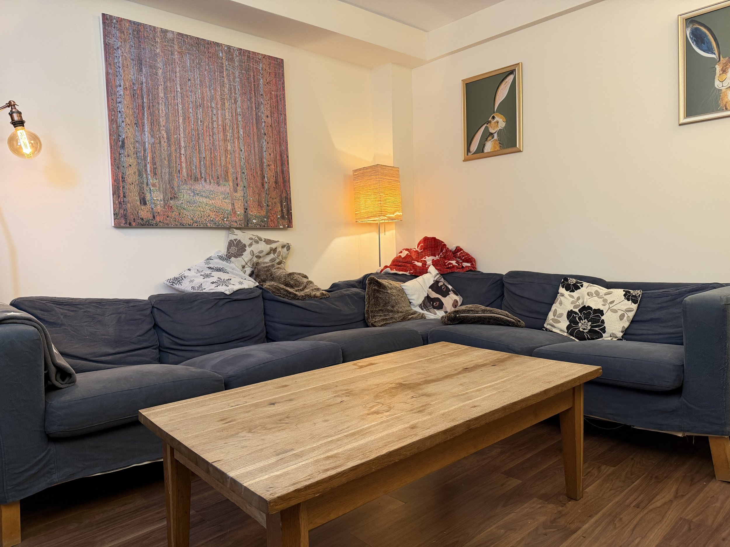 A living room with a large navy blue sectional sofa, multiple pillows, and blankets. There is a wooden coffee table in front of the sofa, a tall floor lamp emitting warm light, and wall art including a large painting of a wooded scene and smaller framed pictures of rabbits with long ears.