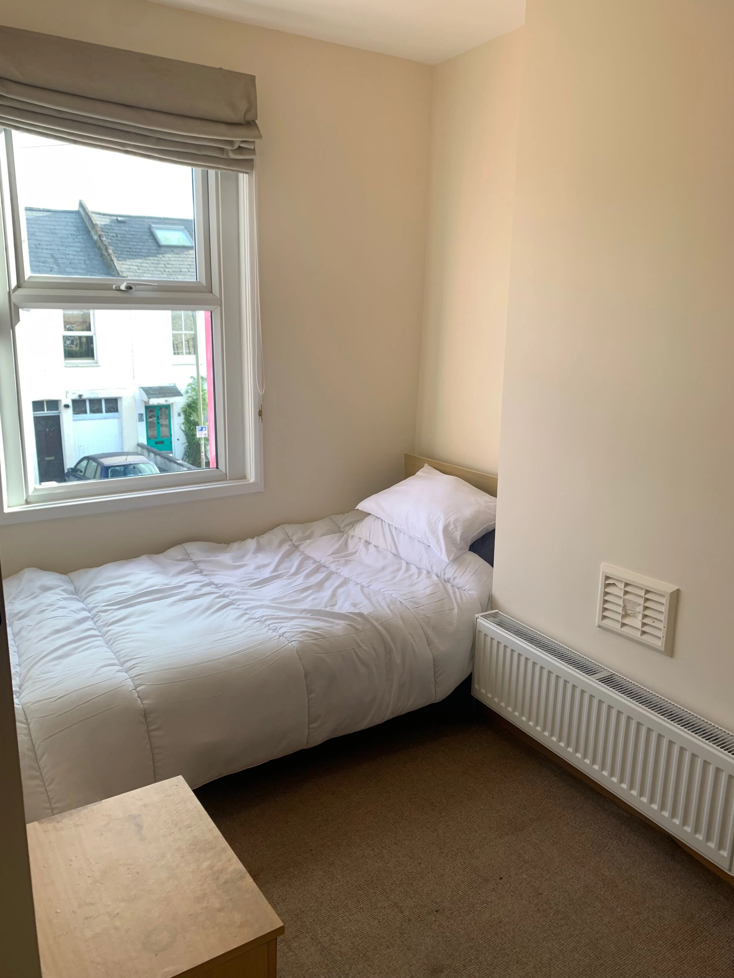 A small bedroom with a single bed, white bedding, and a pillow. There is a window with overlooking neighboring houses and a parked car outside. A radiator and an air vent are on the wall next to the bed, and a wooden nightstand is at the foot of the 