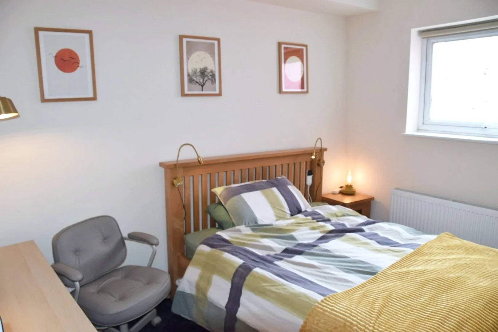 A bedroom with a wooden bed frame and colorful striped bedding, three framed art prints on the wall, a gray office chair, a bedside table with a lamp and a small light, a window with blinds, and a radiator.