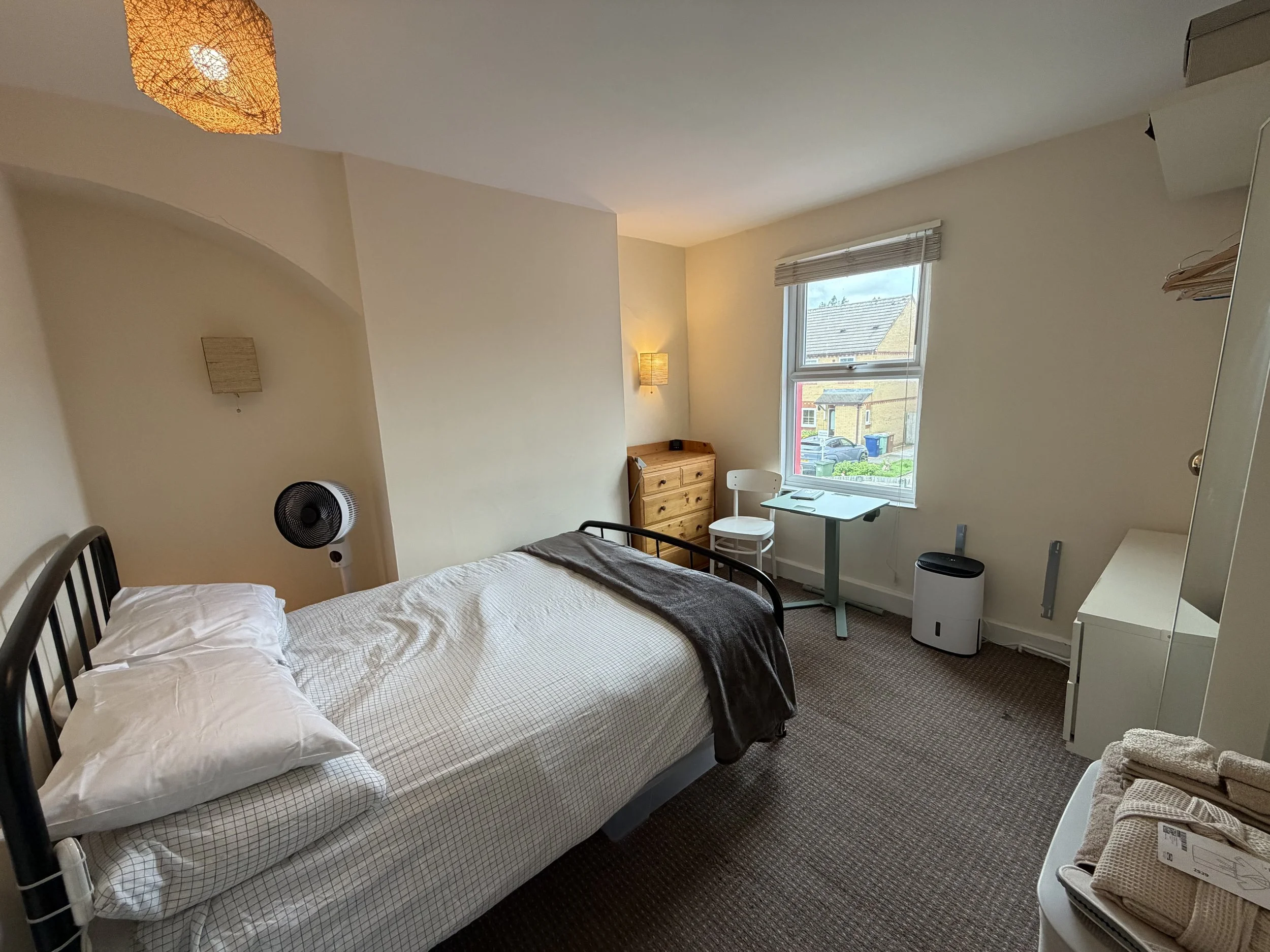 The image shows a tidy bedroom with a single bed, a window with a view of a residential neighborhood, a small wooden dresser, a white chair, a desk, a floor fan, a waste bin, and some shelves. The room has beige walls, woven wall decor, and a ceiling light fixture.