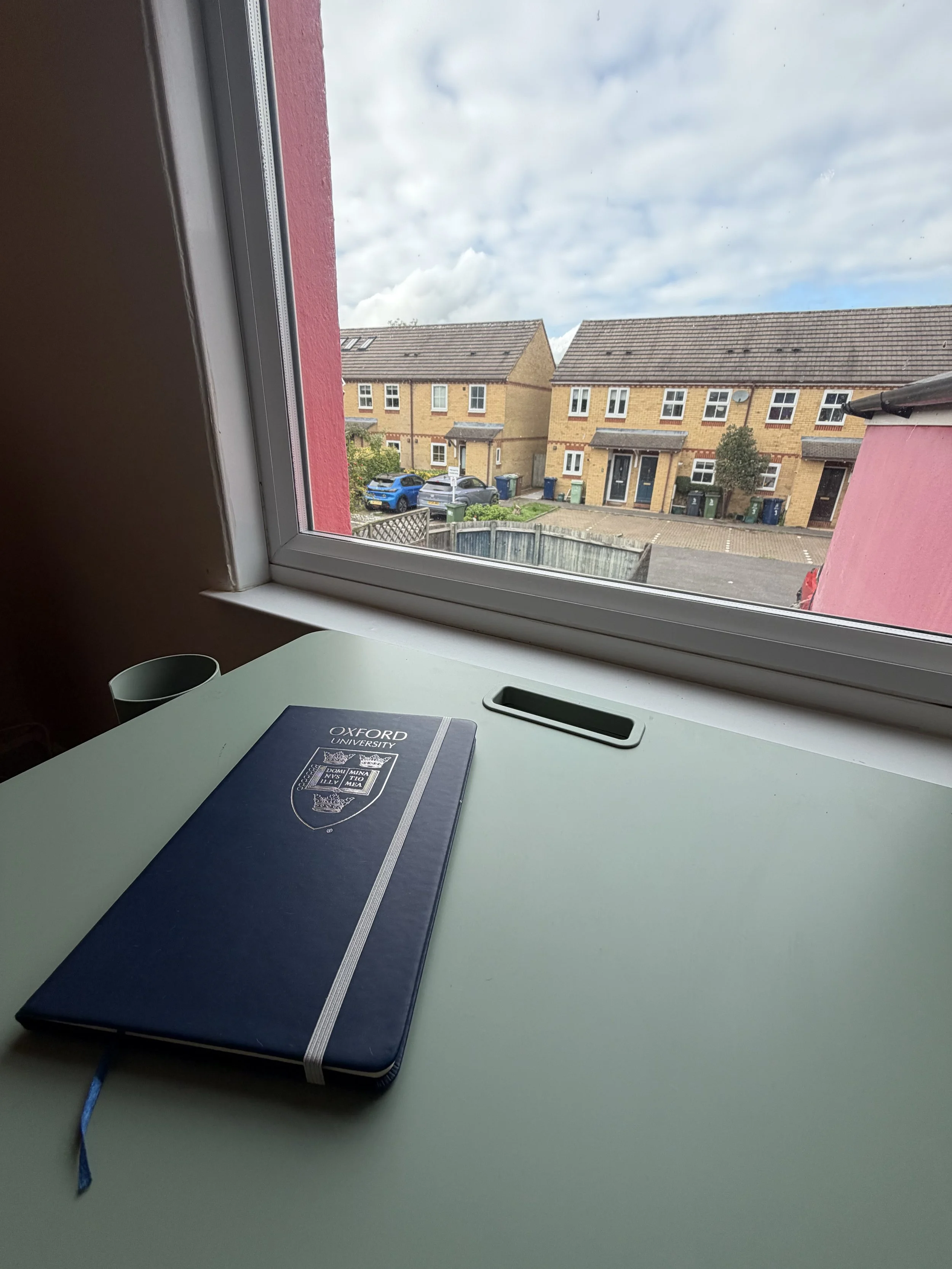 View from an indoor desk shows a notebook labeled 'Oxford University' on a white desk, with a window overlooking a residential street with houses, parked cars, and a cloudy sky.