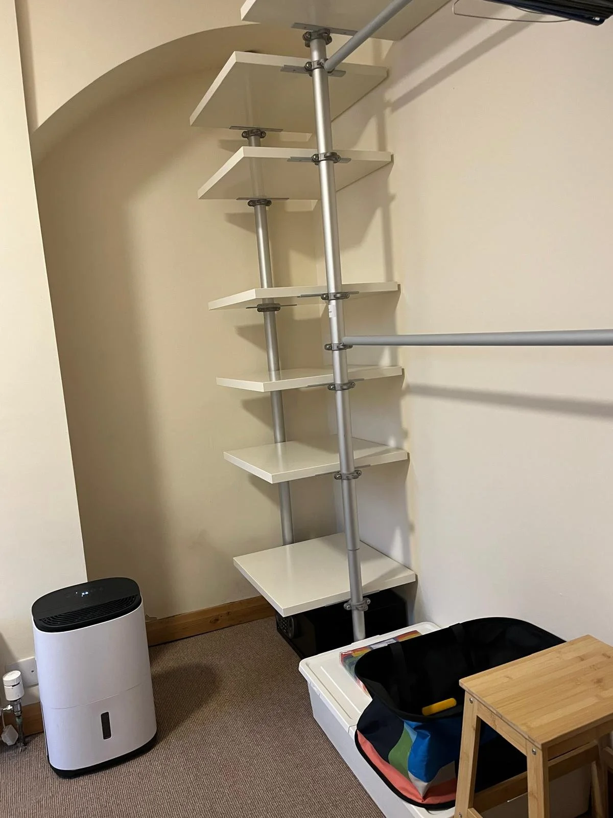 A corner of a room with a stepladder with white shelves and gray metal supports against a cream-colored wall. A white air purifier, a multicolored bag, and a small wooden stool are on the beige carpet floor.