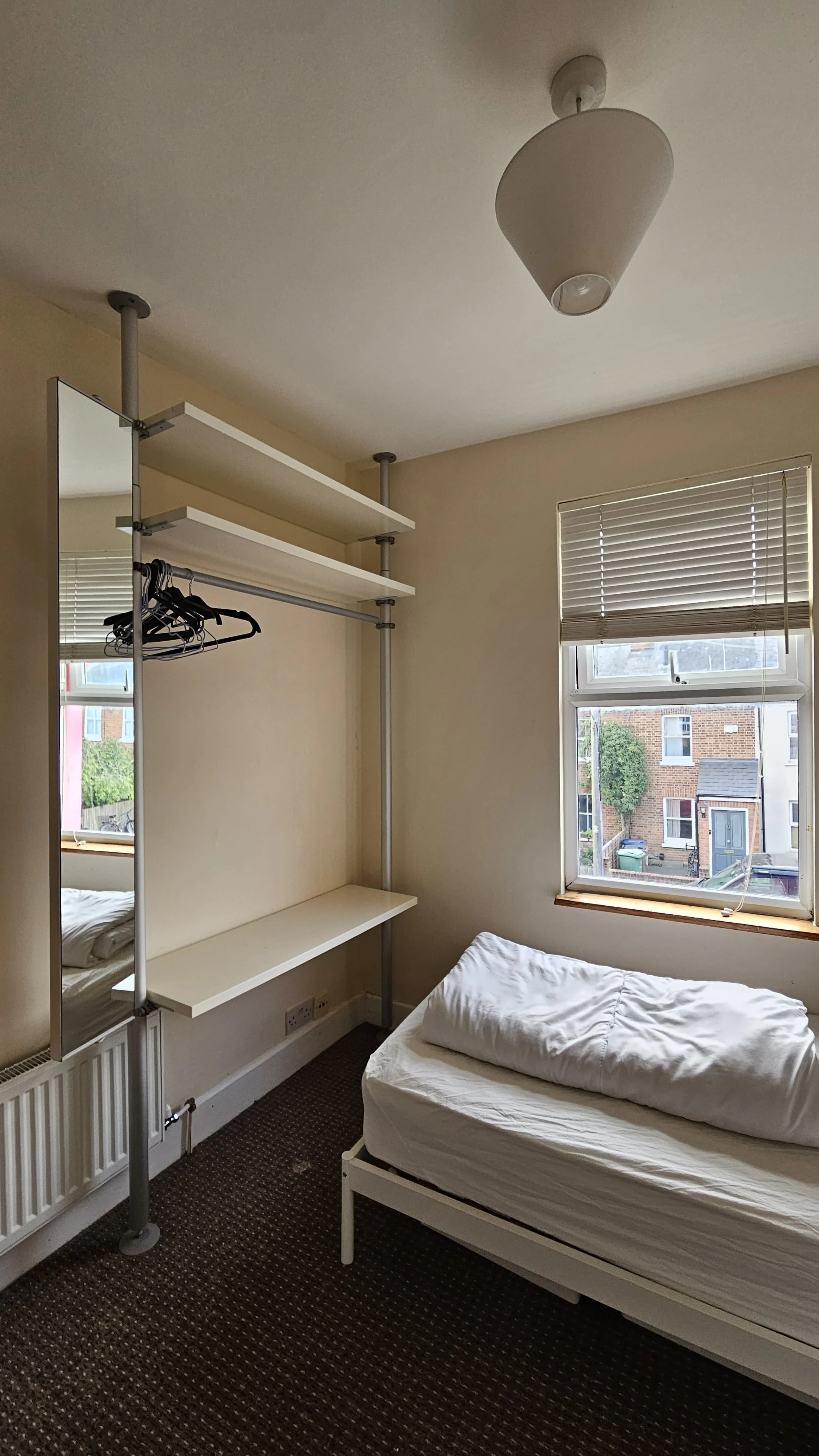 A small, sparsely furnished bedroom with a single bed, a window with blinds, and a basic clothing rack with a mirror.
