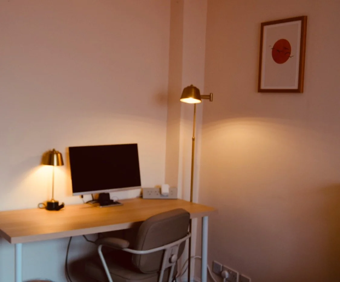 A home office corner with a desk, a computer, a lamp, and a picture of a smiling sun on the wall.