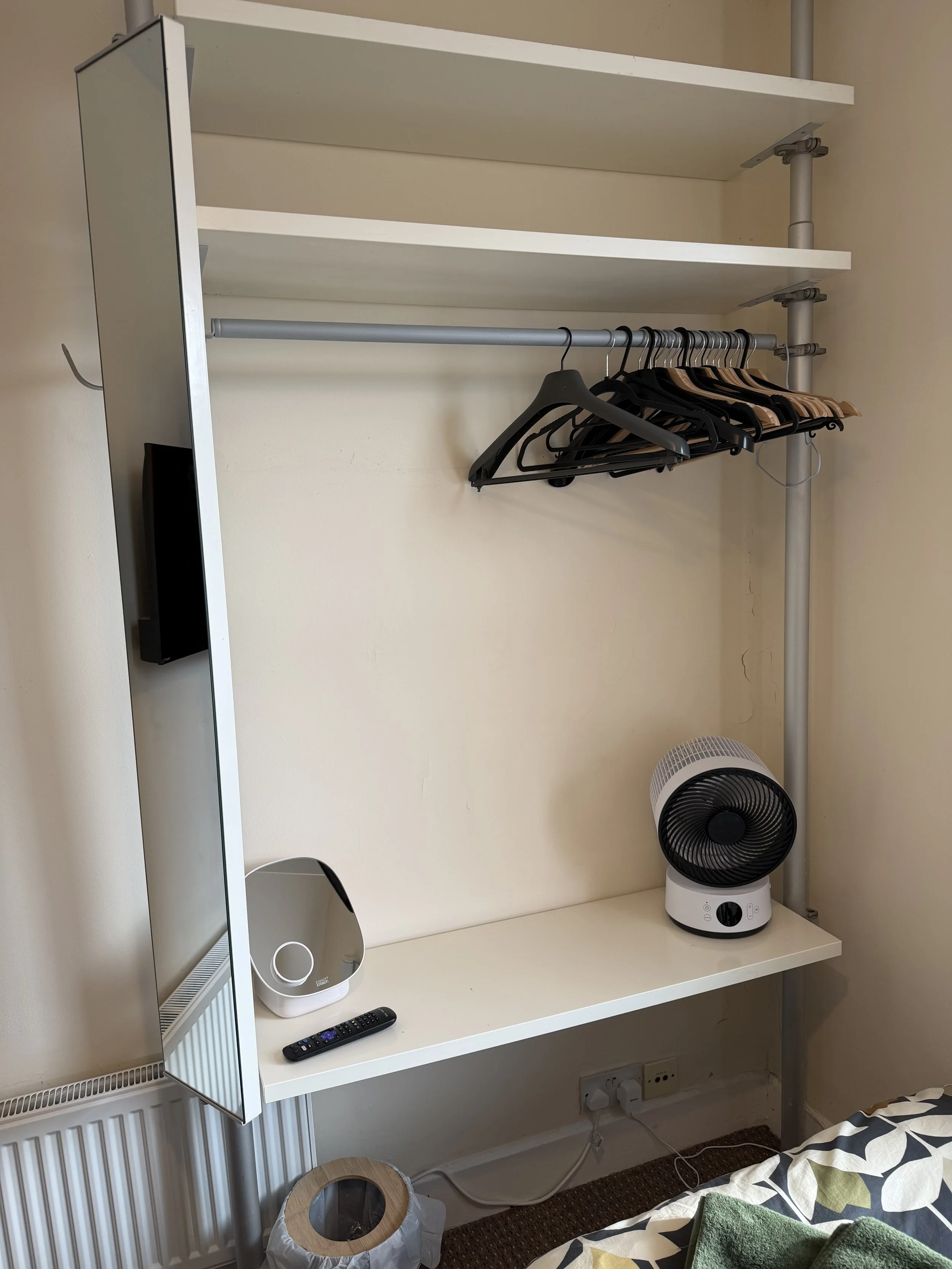 Empty clothing rack with a few black and wooden hangers, a small mirror, a fan, a remote control, and a compact heater on a white shelf in a room.