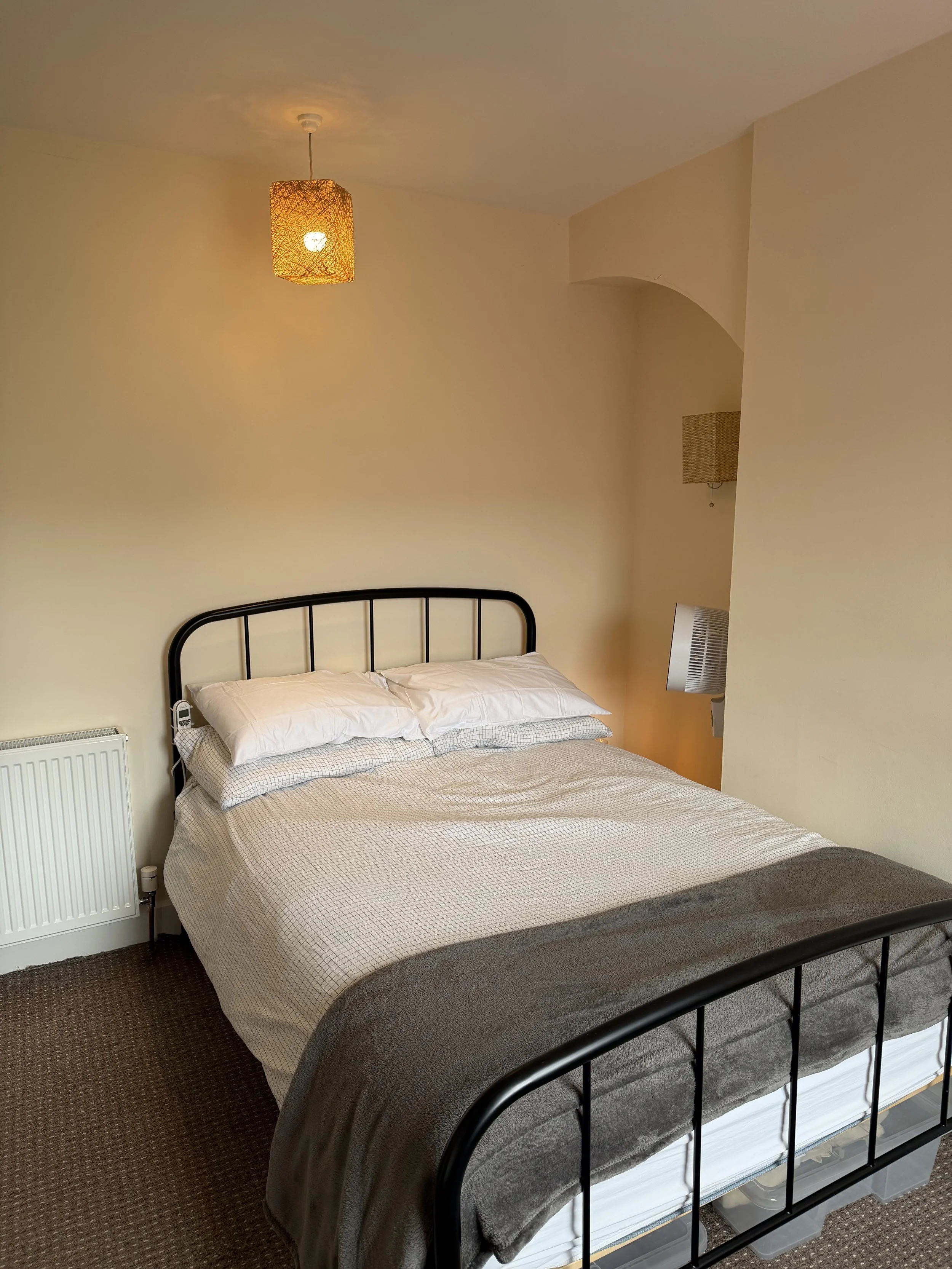A neatly made bed with white pillows and a light-colored checked duvet in a cozy bedroom. There is a black metal headboard, a radiator, a small side table with a lamp, and a ceiling light fixture with a woven lampshade.