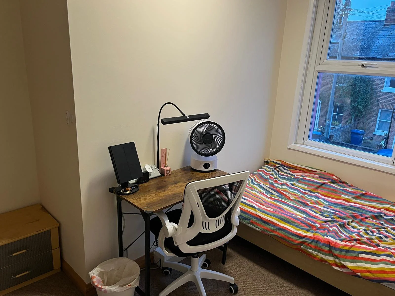 Small bedroom with a bed covered in a colorful striped blanket, a desk with a black monitor, a fan, and a white office chair, beside a window showing an urban street scene.