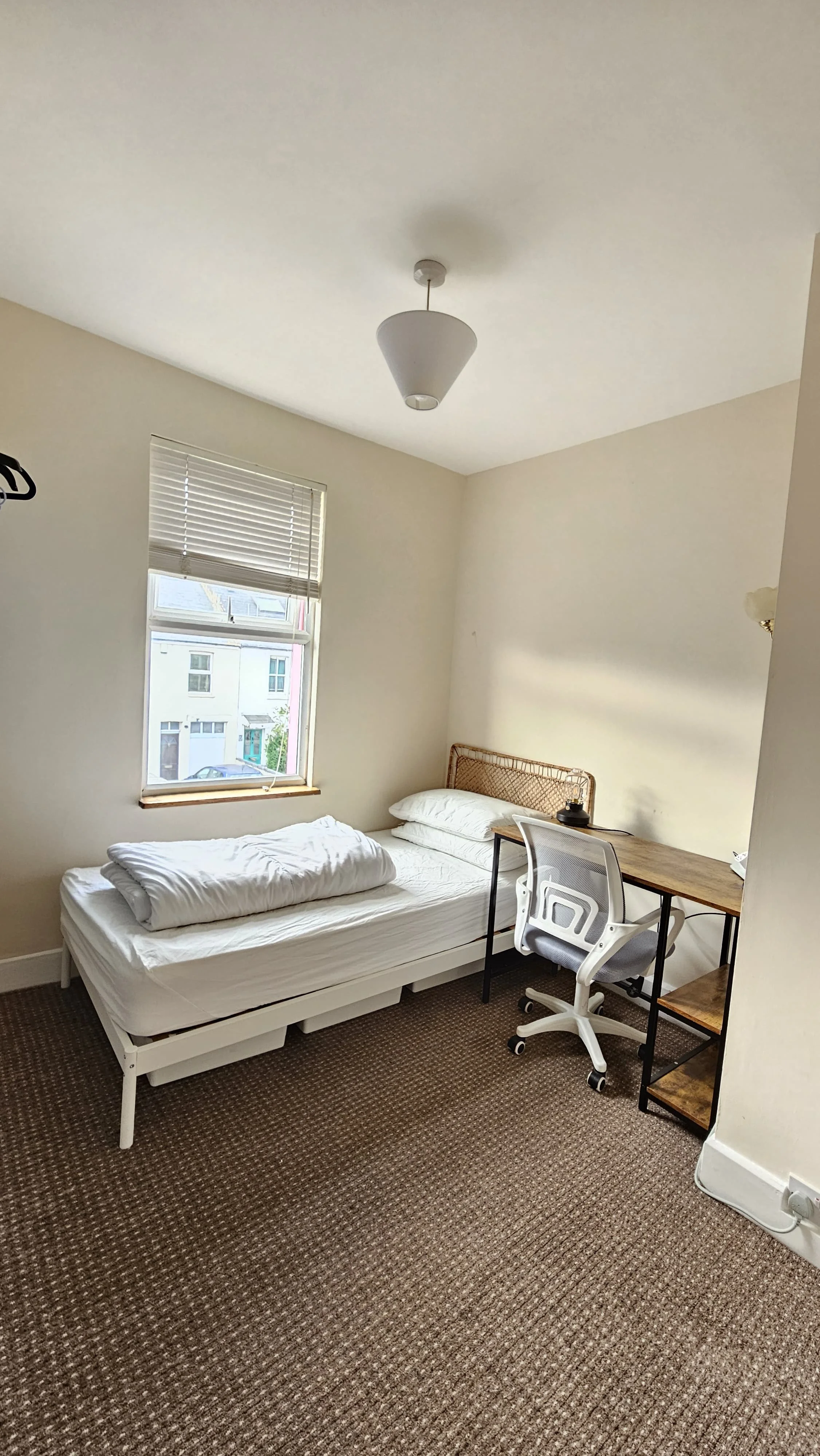A simple bedroom with a single bed, a window with blinds, a wooden desk with a lamp, a white office chair, and brown patterned carpet.