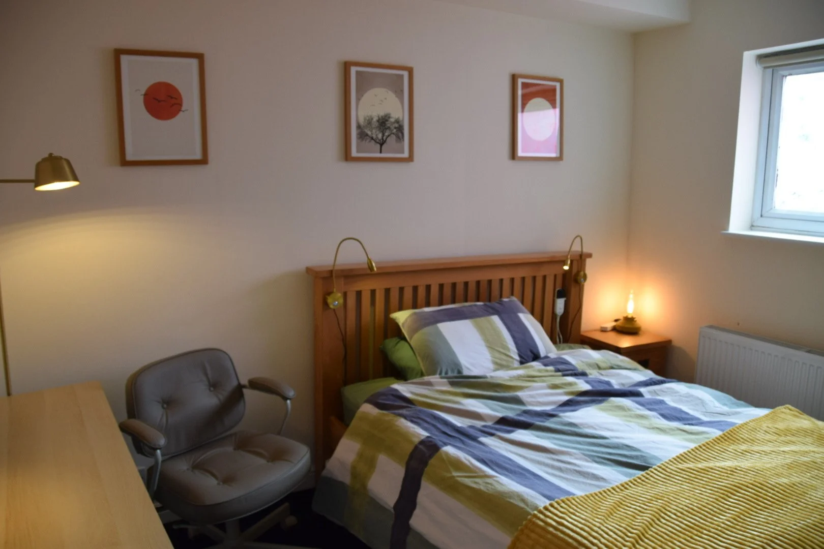 A cozy bedroom with a wooden bed frame, plaid bedding, and colorful artwork on the wall, illuminated by soft lighting.