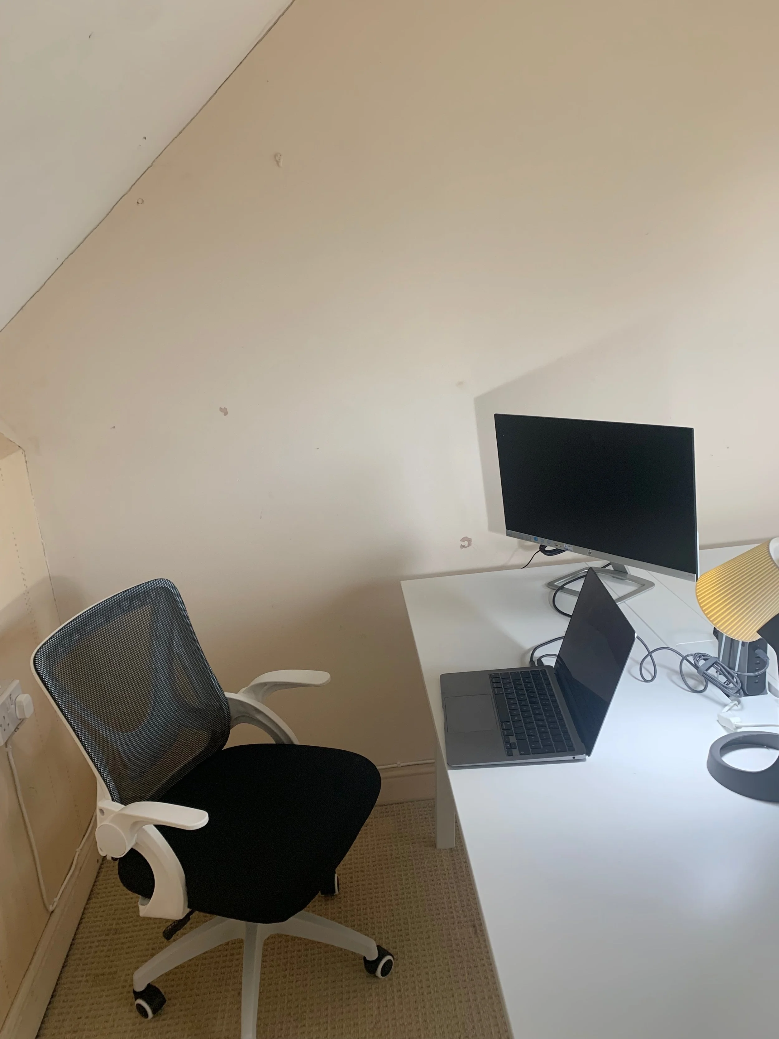 A small home office with a white desk, a black office chair, a laptop, a monitor, a yellow lamp, and a beige carpet.