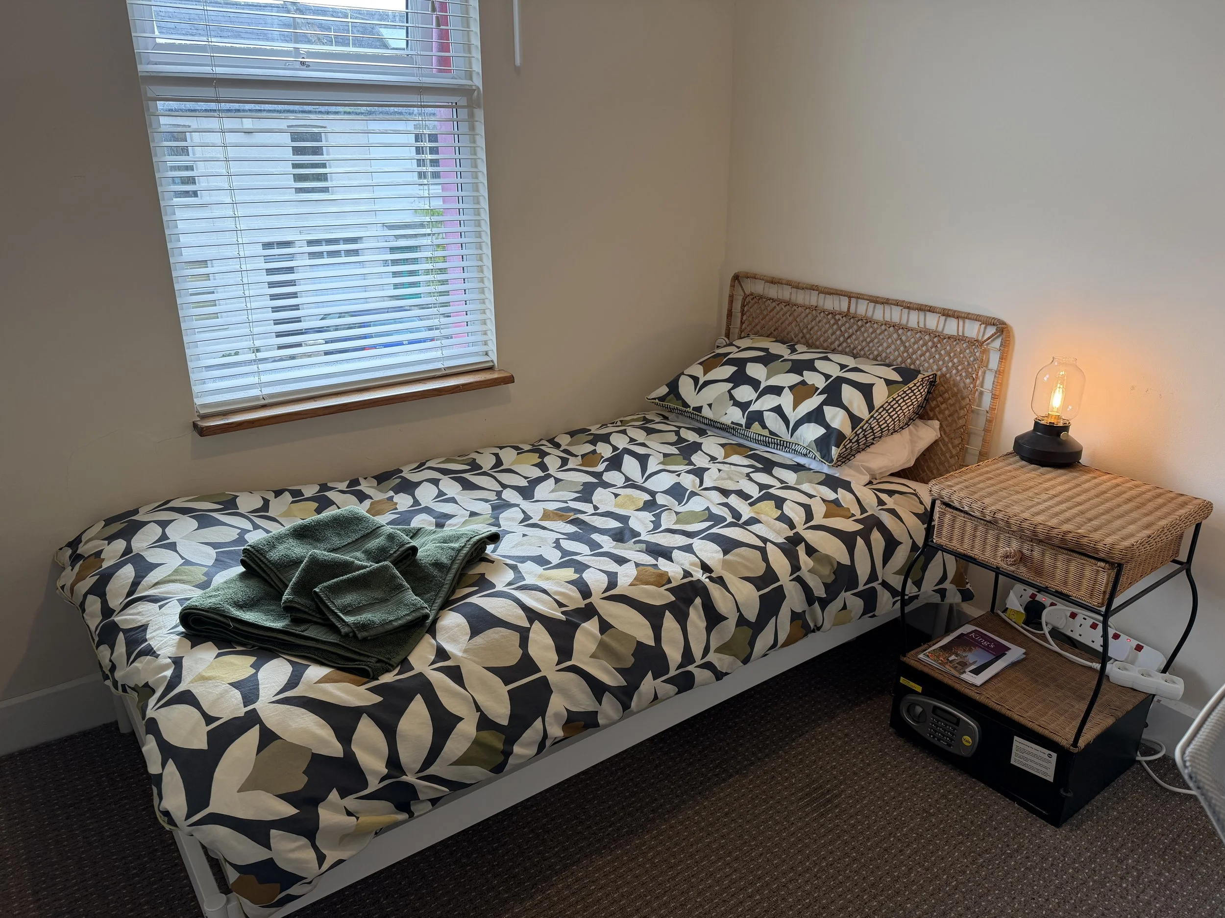 A tidy single bed with a patterned duvet and a matching pillow, green towels folded at the foot, a window with white blinds near the bed, a small wicker nightstand with a lamp, magazine, and electrical outlet