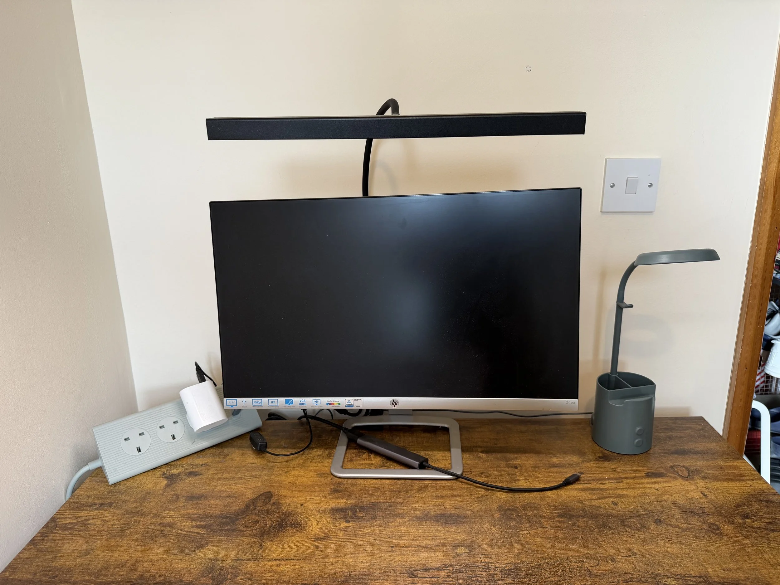 A computer monitor on a wooden desk with a black strip light above, a gray desk lamp to the right, and a white power strip with outlets to the left.