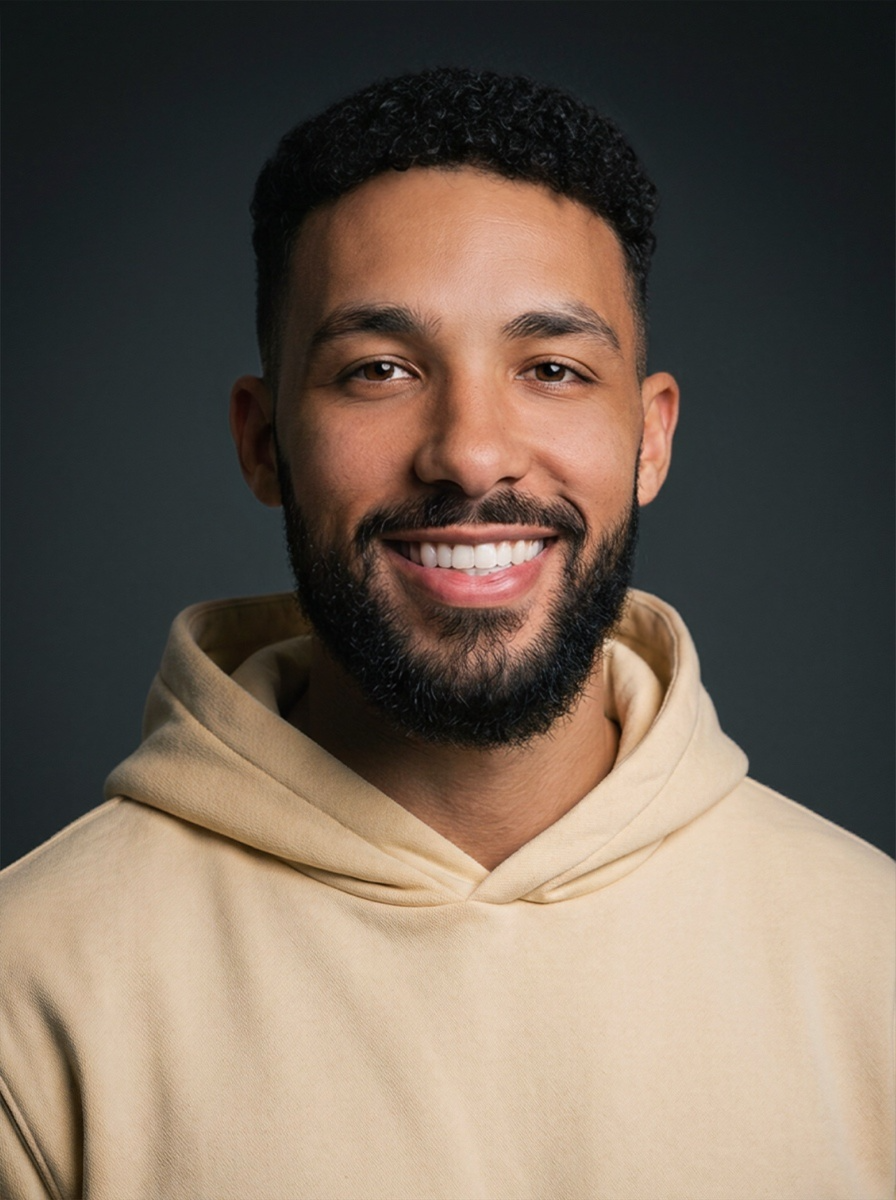 Portrait of a smiling man with short curly hair and a beard, wearing a beige hoodie against a dark background.