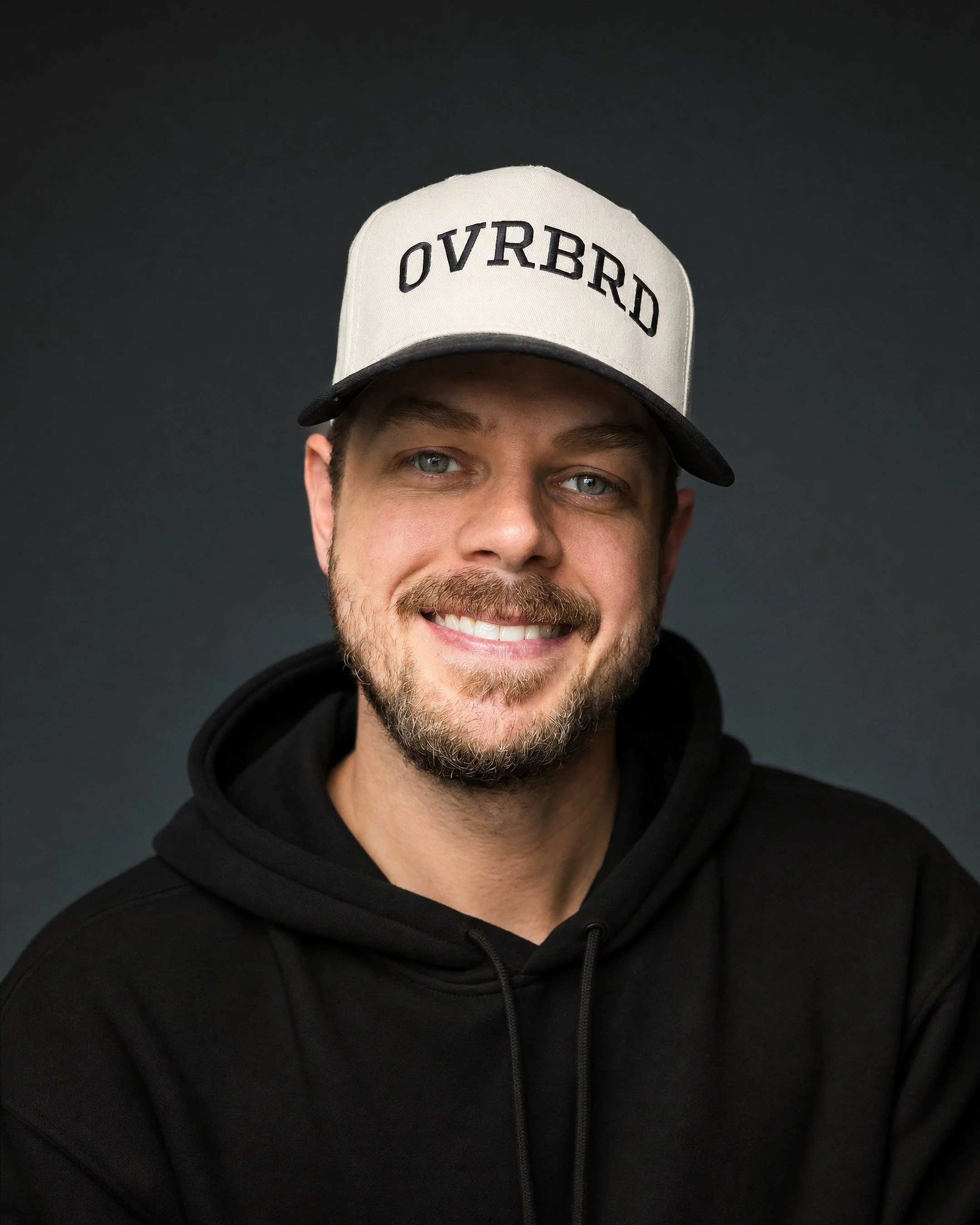 Portrait of a young man with dark hair, beard, and mustache, smiling, wearing a black hoodie, against a dark background.