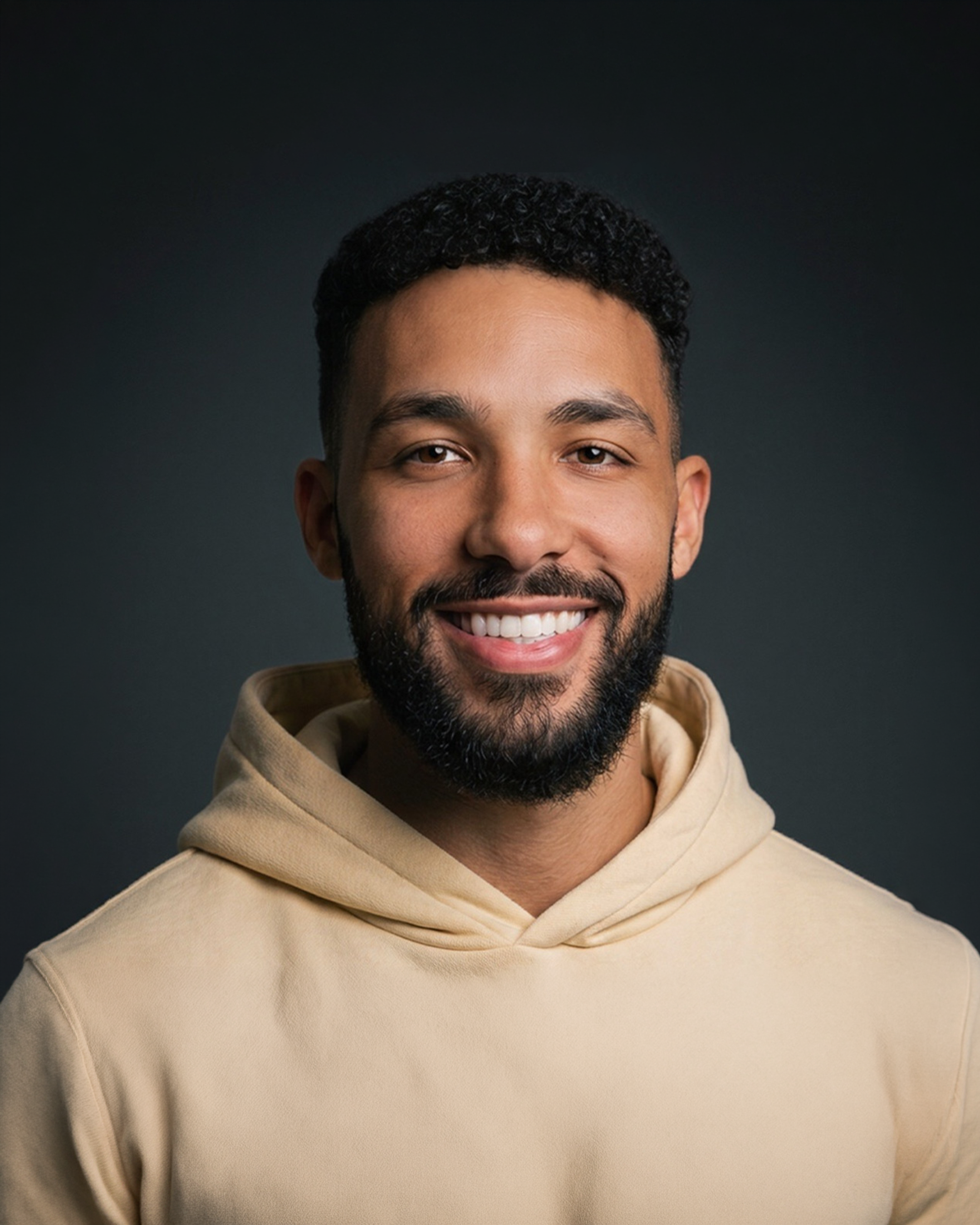 Portrait of a smiling man with short curly hair and a beard, wearing a beige hoodie against a dark background.