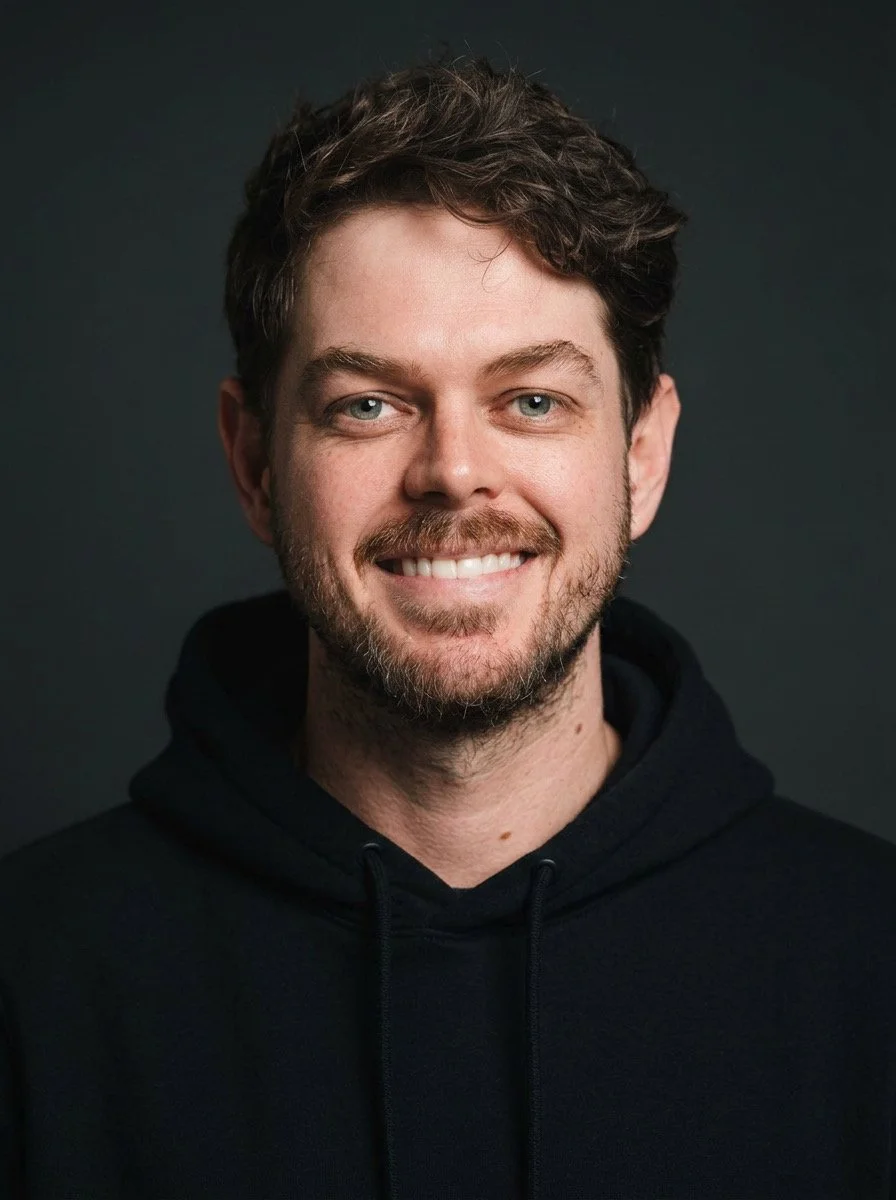 Portrait of a young man with dark hair, beard, and mustache, smiling, wearing a black hoodie, against a dark background.