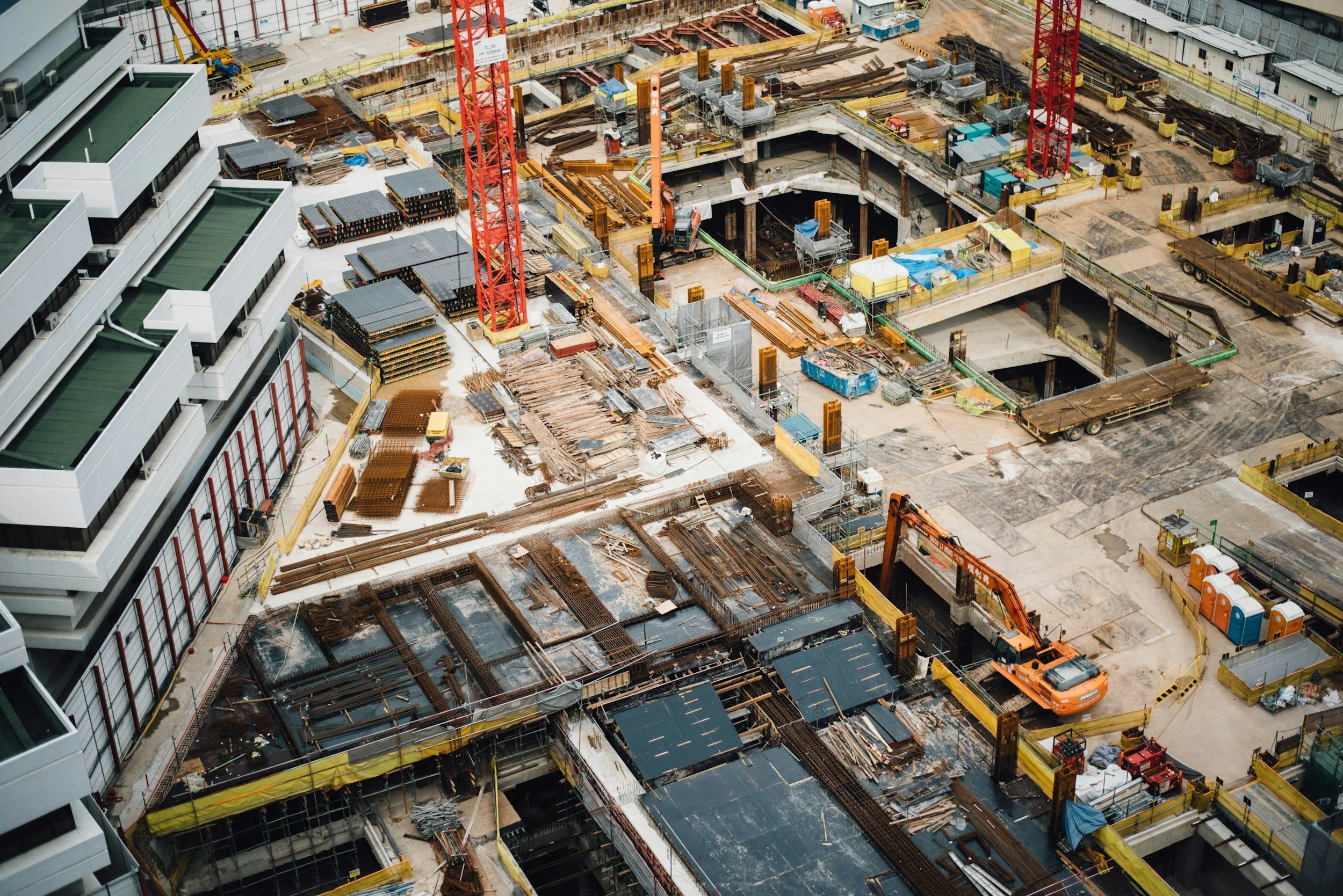 Aerial view of a construction site with multiple deep excavations, building materials, scaffolding, and construction equipment including cranes and an orange excavator. Part of an adjacent modern white building with green roofs is visible on the left.
