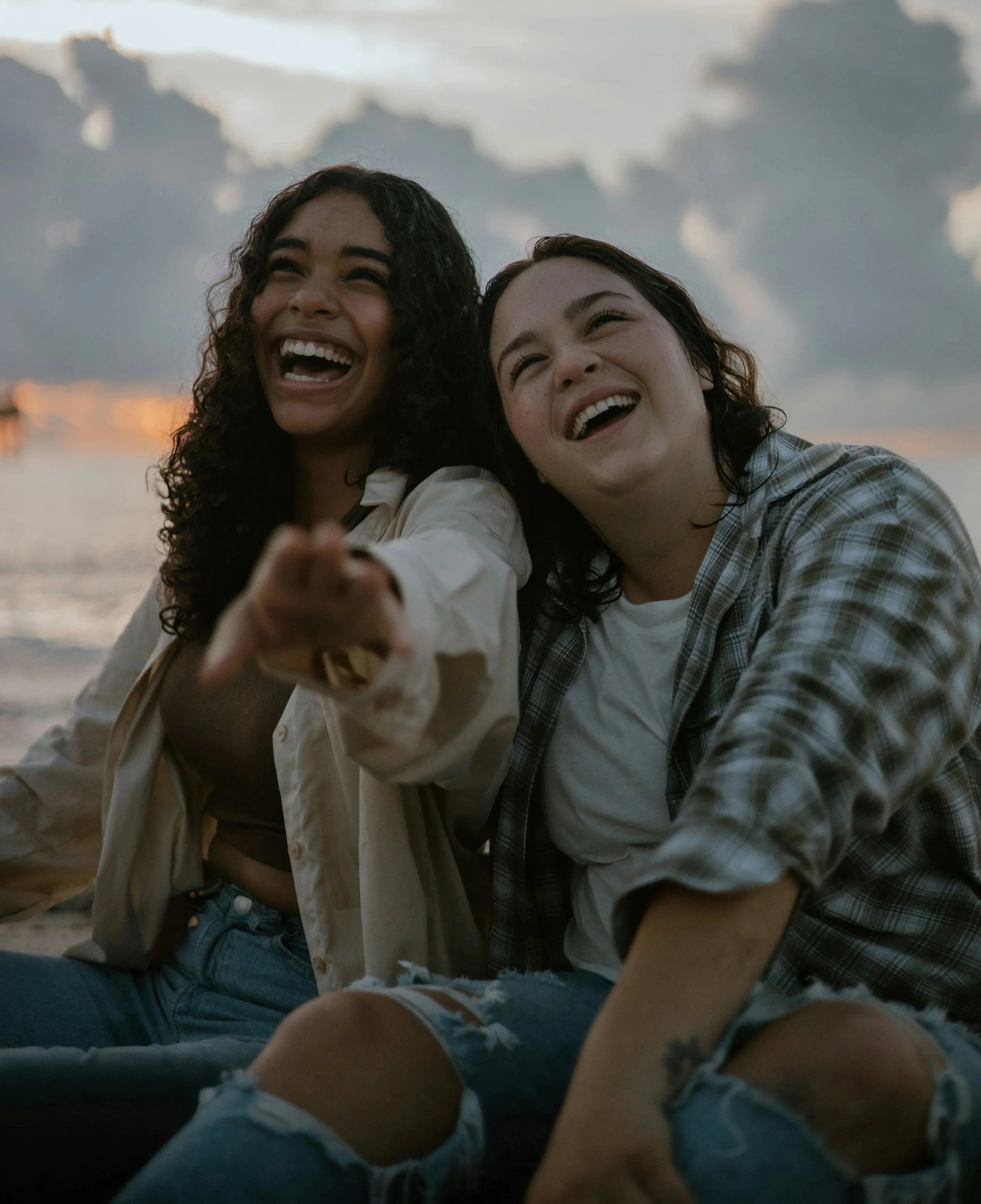 Two women sitting on the beach, smiling and enjoying each other's company during sunset.