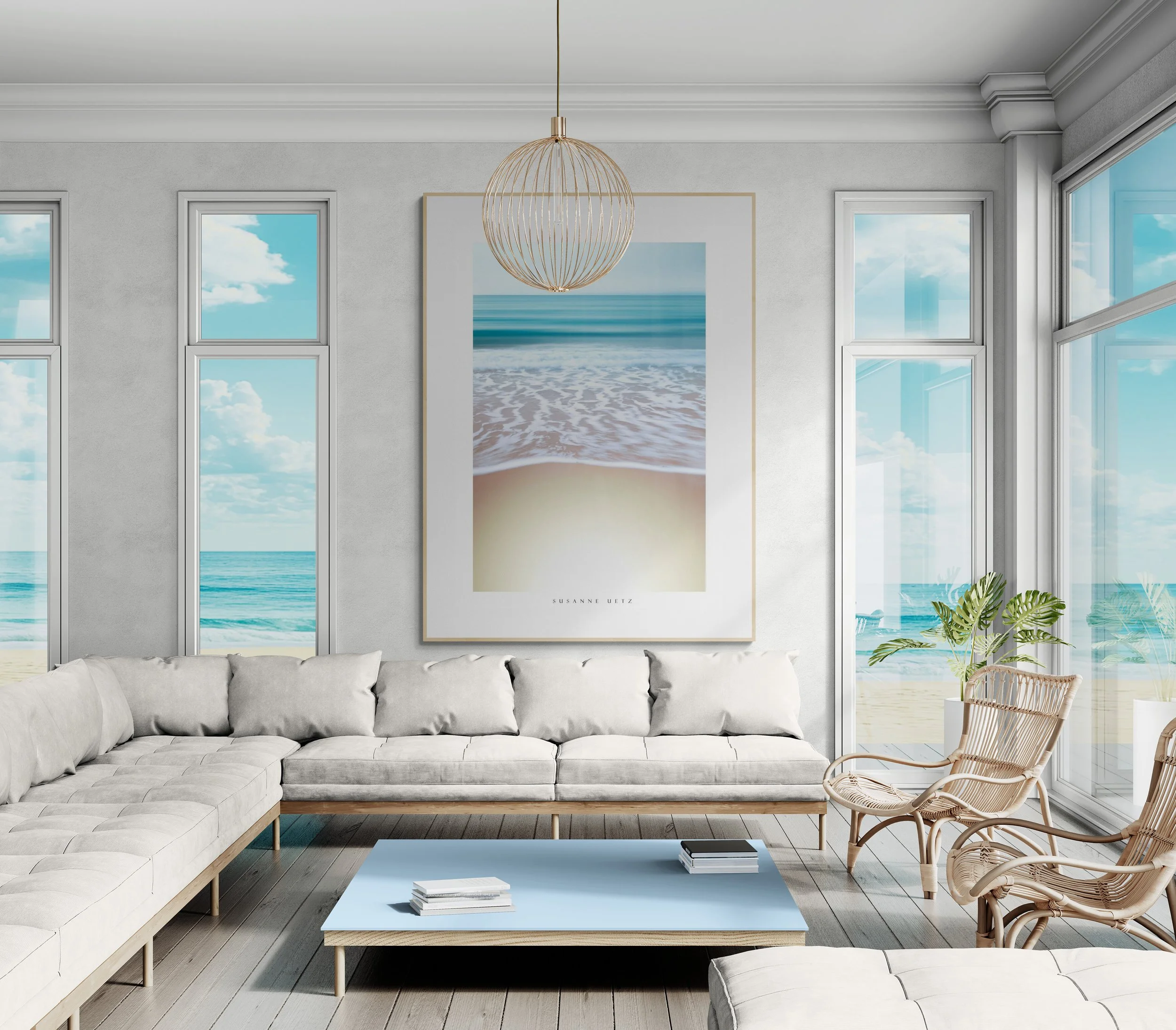 Modern living room with large windows overlooking the beach, white sofa, wicker chairs, a light blue coffee table with books, and a large beach-themed artwork on the wall.