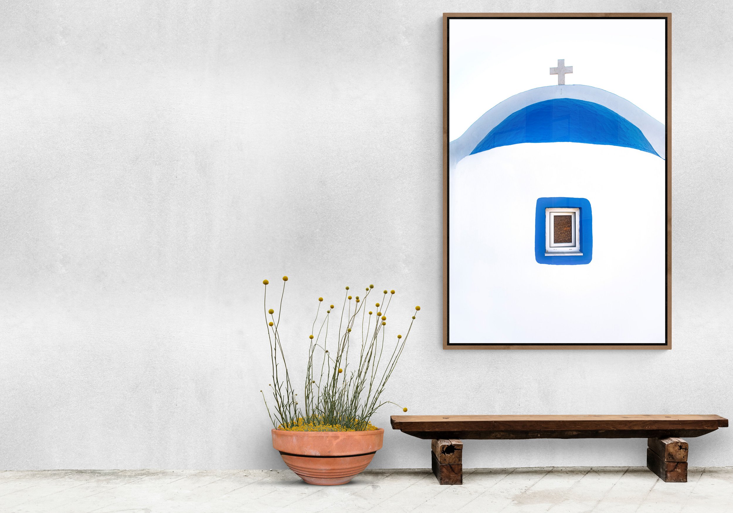 Interior of a room with a white wall, a framed picture of a blue-domed building with a cross on top, a wooden bench, and a potted plant with yellow flowers.