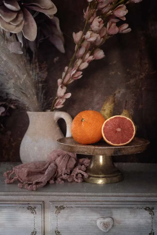 A still life arrangement with a vase holding pink flowers, a whole orange, and a halved grapefruit with pink flesh, placed on a wooden cake stand. A pink cloth is draped on the surface below.