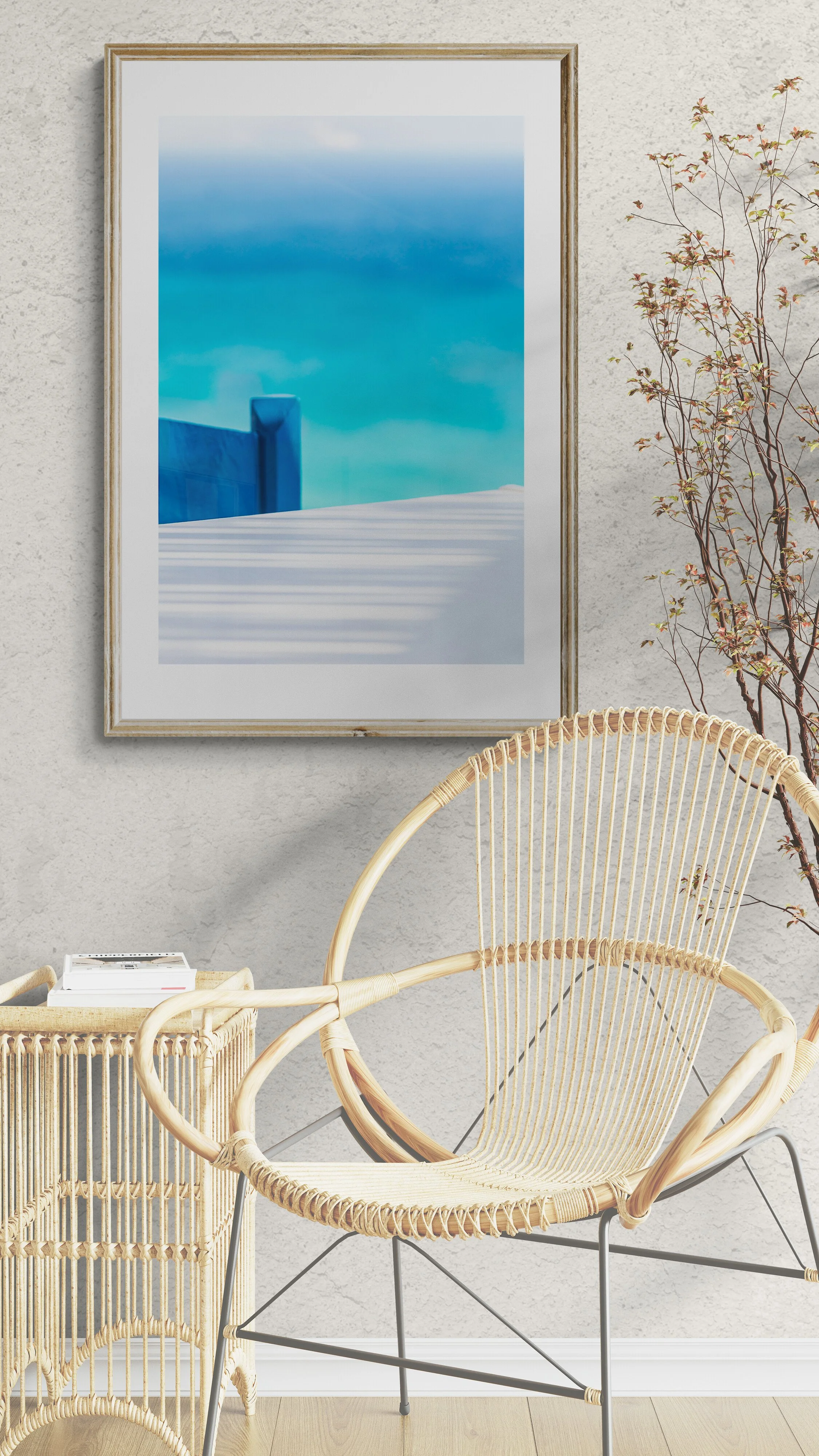 Interior scene with a wicker chair, a side table with books, and a framed picture of a blue and white landscape on the wall, with a decorative branch in the background.