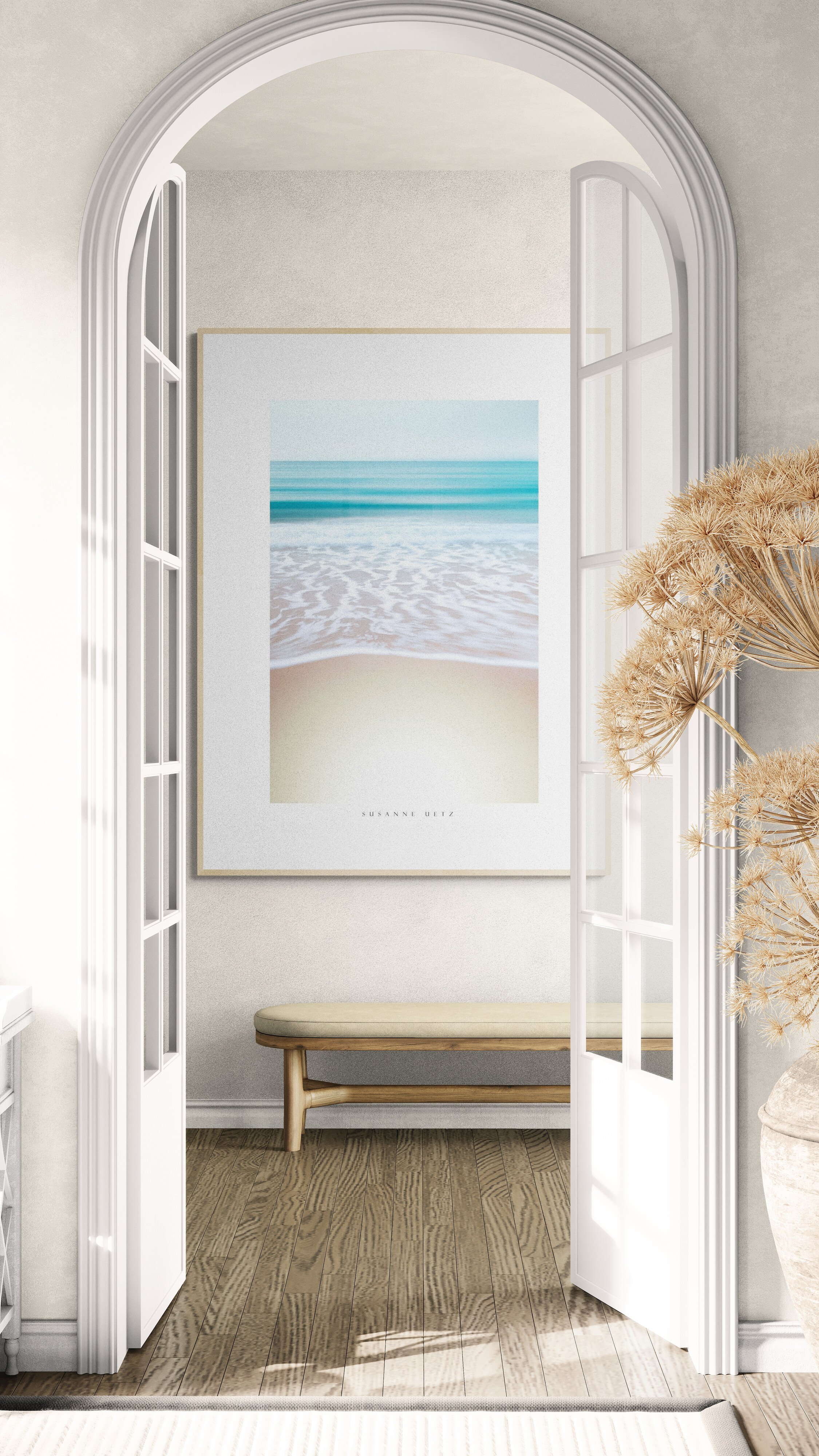 Interior view through white French doors showing a framed beach scene print on a wall with a wooden bench underneath and beige dried floral arrangement on the right.
