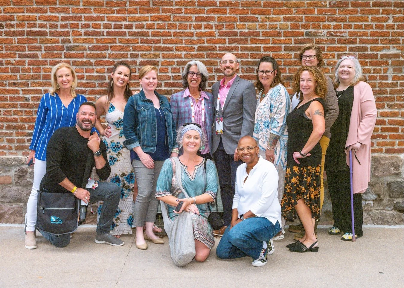Professional Event Photography by Minneapolis Photographer Jay Cupcake. A group of fifteen diverse adults posing for a photo against a brick wall, smiling and dressed casually.