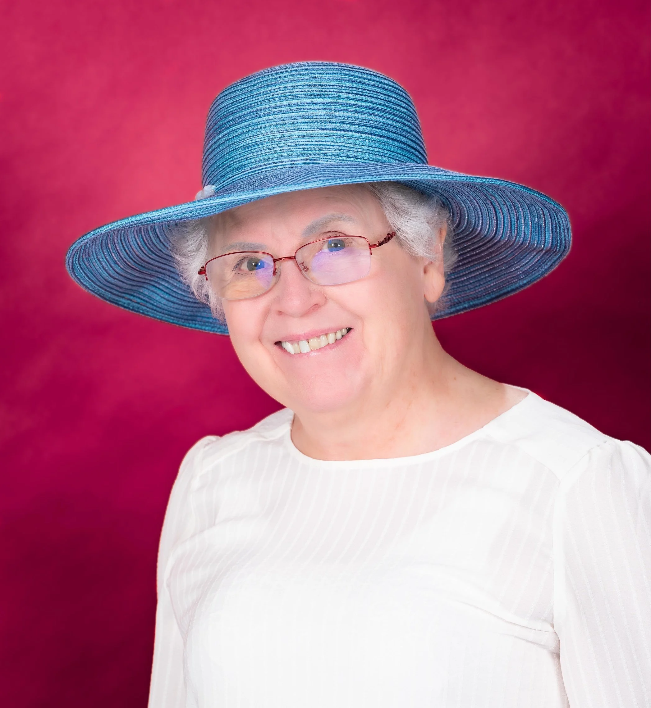 Professional Portrait Photography by Saint Paul Photographer Jay Cupcake. A smiling older woman wearing a wide-brimmed blue hat, glasses, and a white top against a pink background.