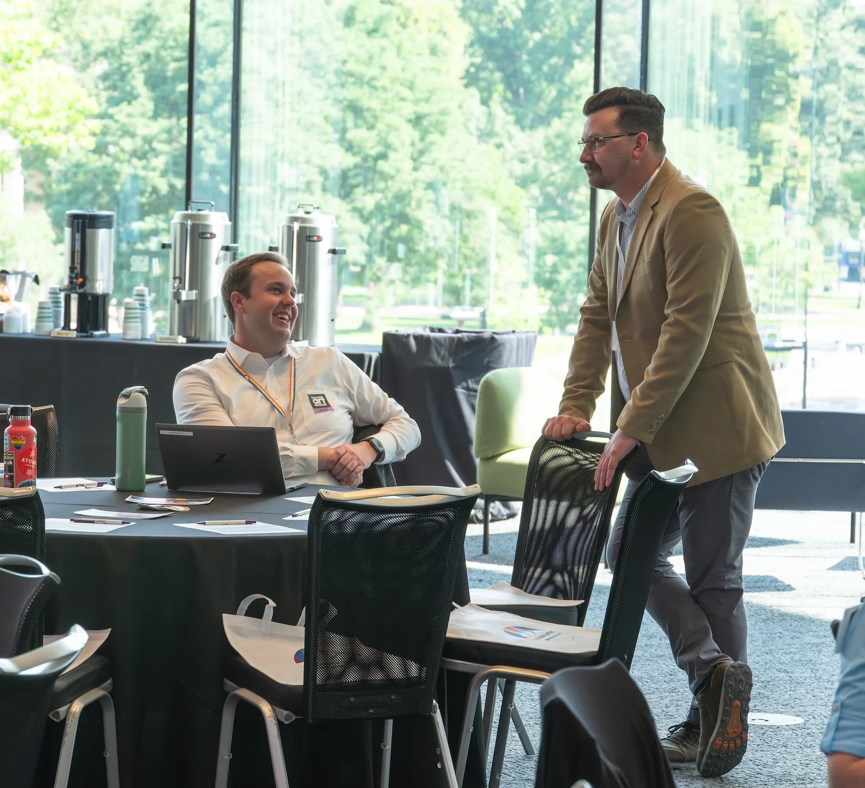 Professional Event Photography by Twin Cities Photographer Jay Cupcake. Two men in a conference room, one sitting and smiling, the other standing and leaning on a chair, engaged in conversation. The room has large windows with outdoor greenery visibl